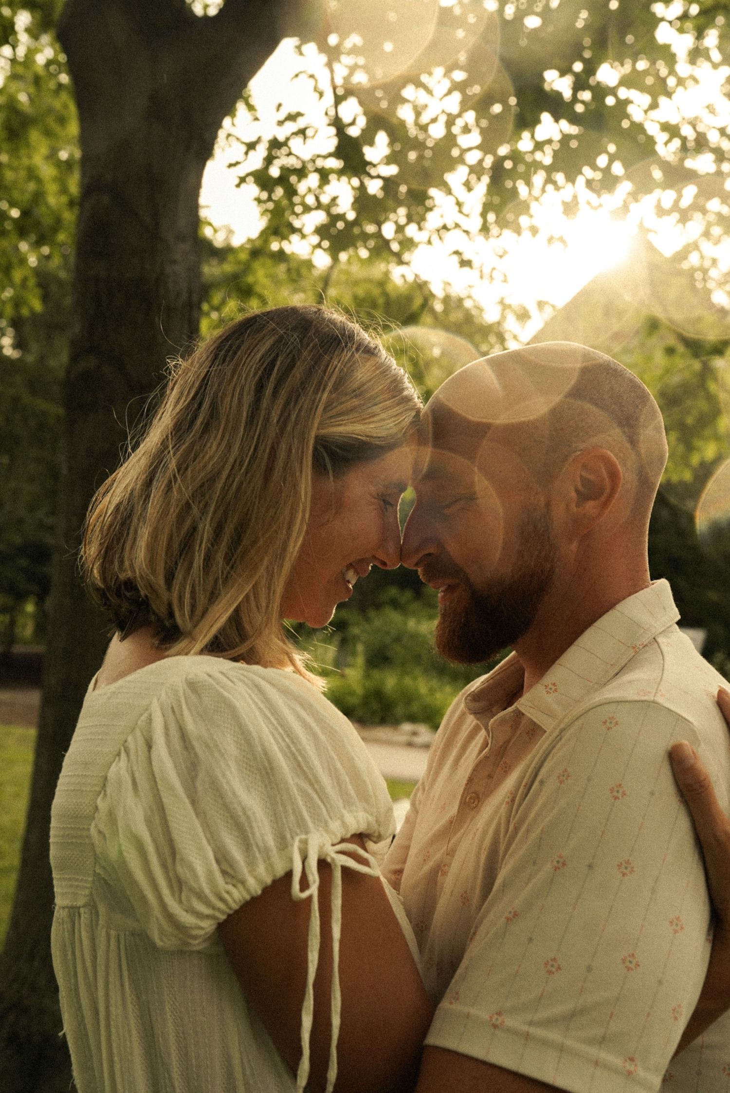 A couple shares a tender forehead-to-forehead moment embracing outdoors in a sunlit park with golden bokeh light.