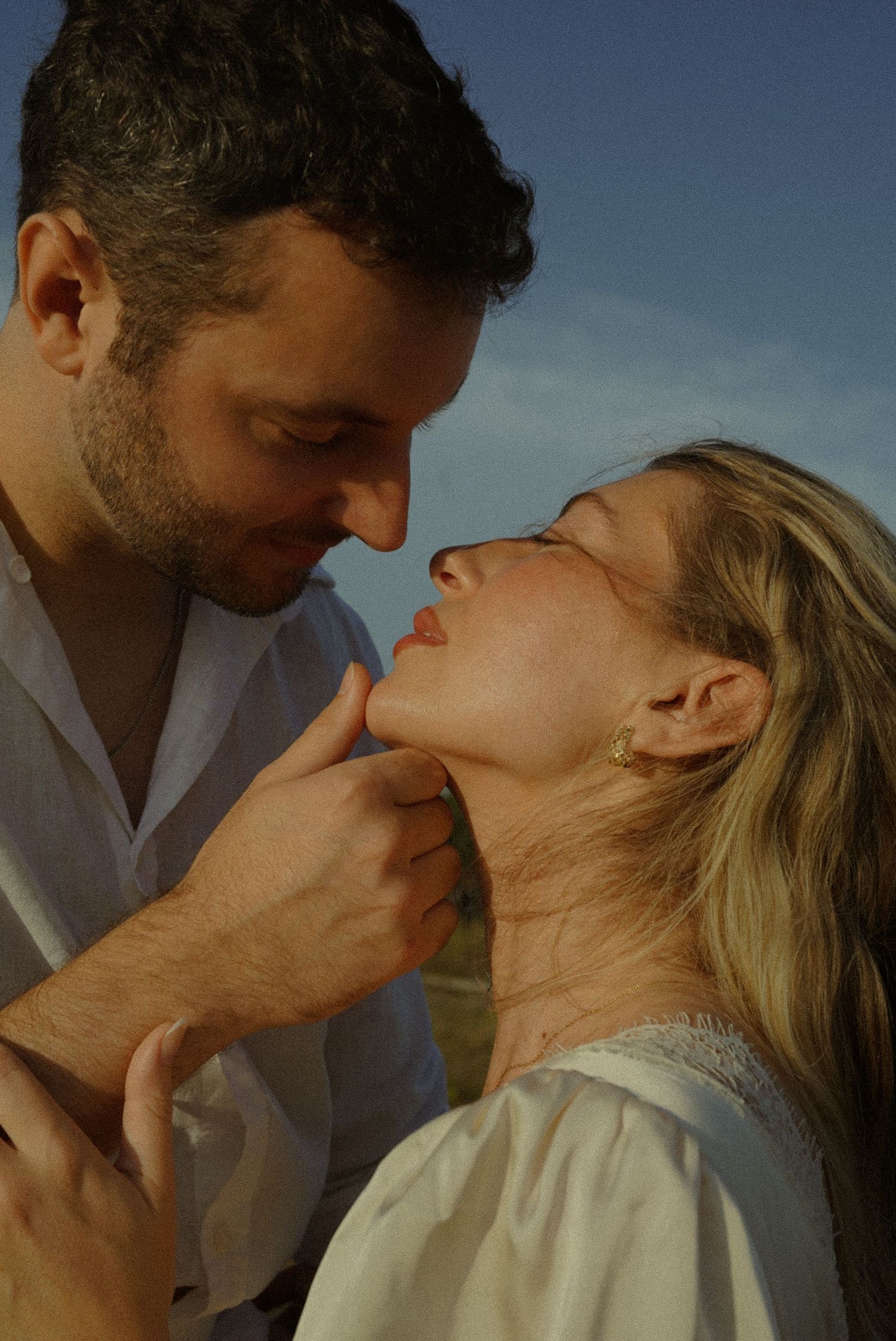 A couple shares a romantic almost-kiss moment outdoors, man gently lifting woman's chin under warm golden light.