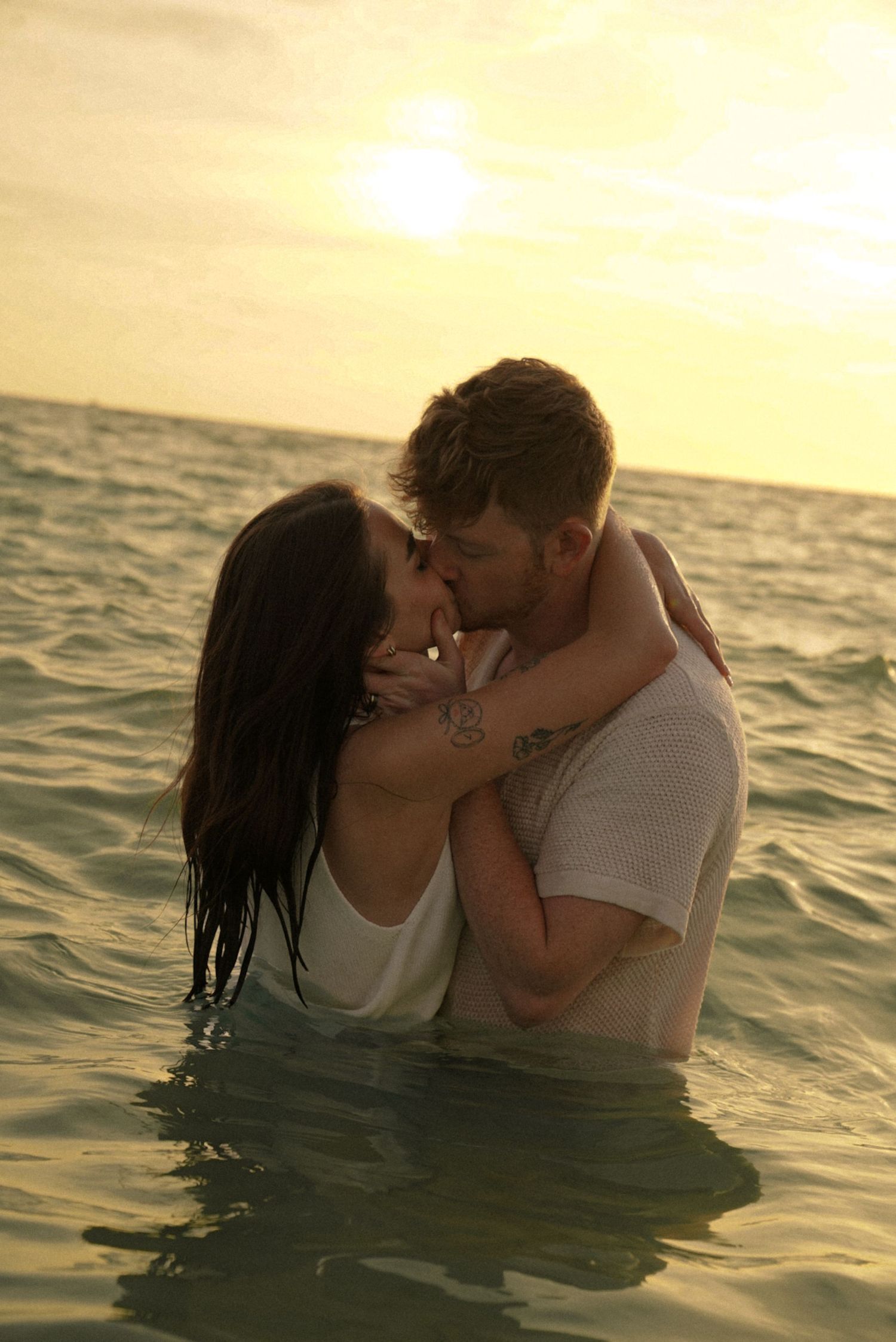 Romantic couple kissing in ocean waves at sunset, standing waist-deep in water with golden sky backdrop.