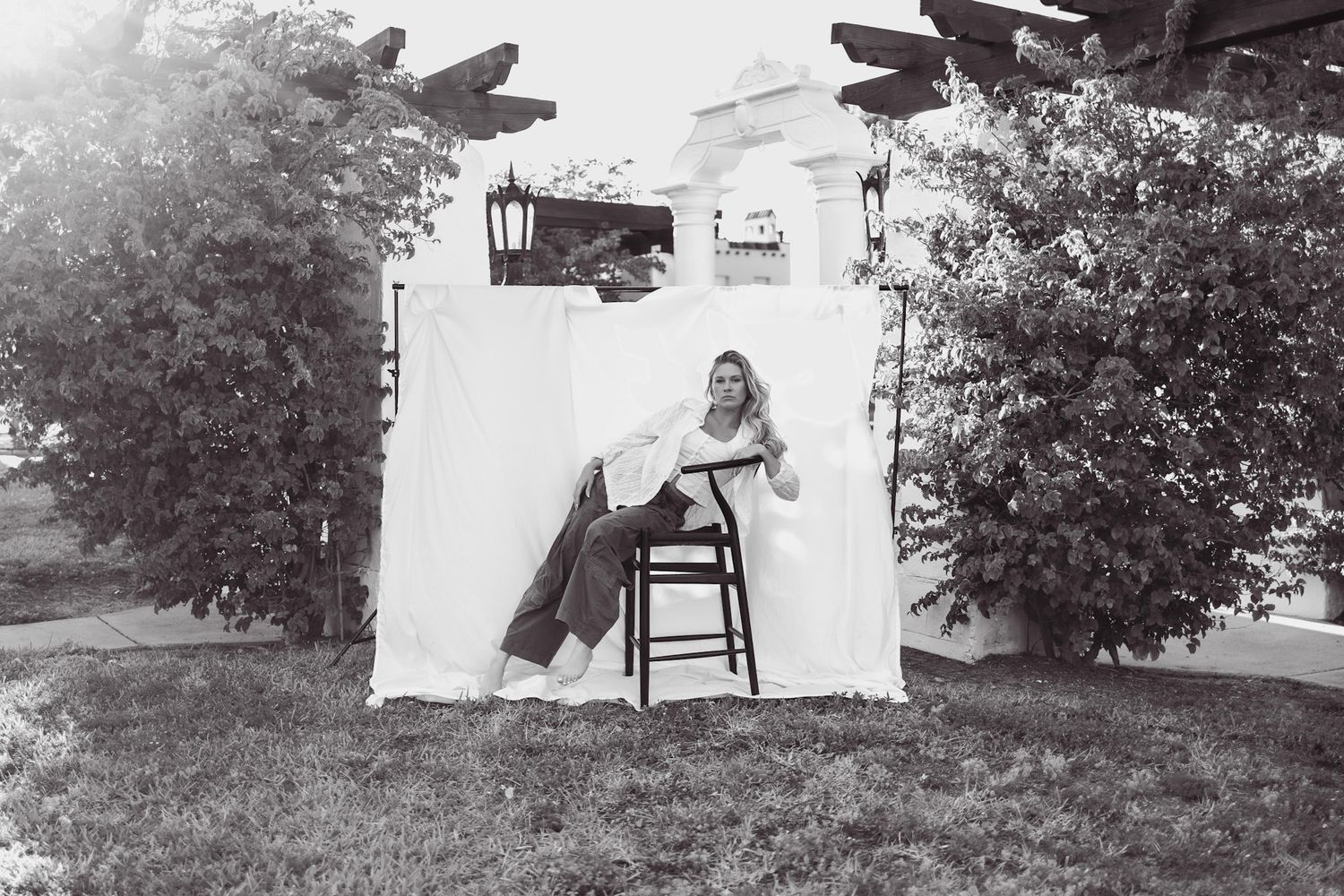 Black and white photo of a woman sitting on a chair outdoors beside a white backdrop near trees and a building.
