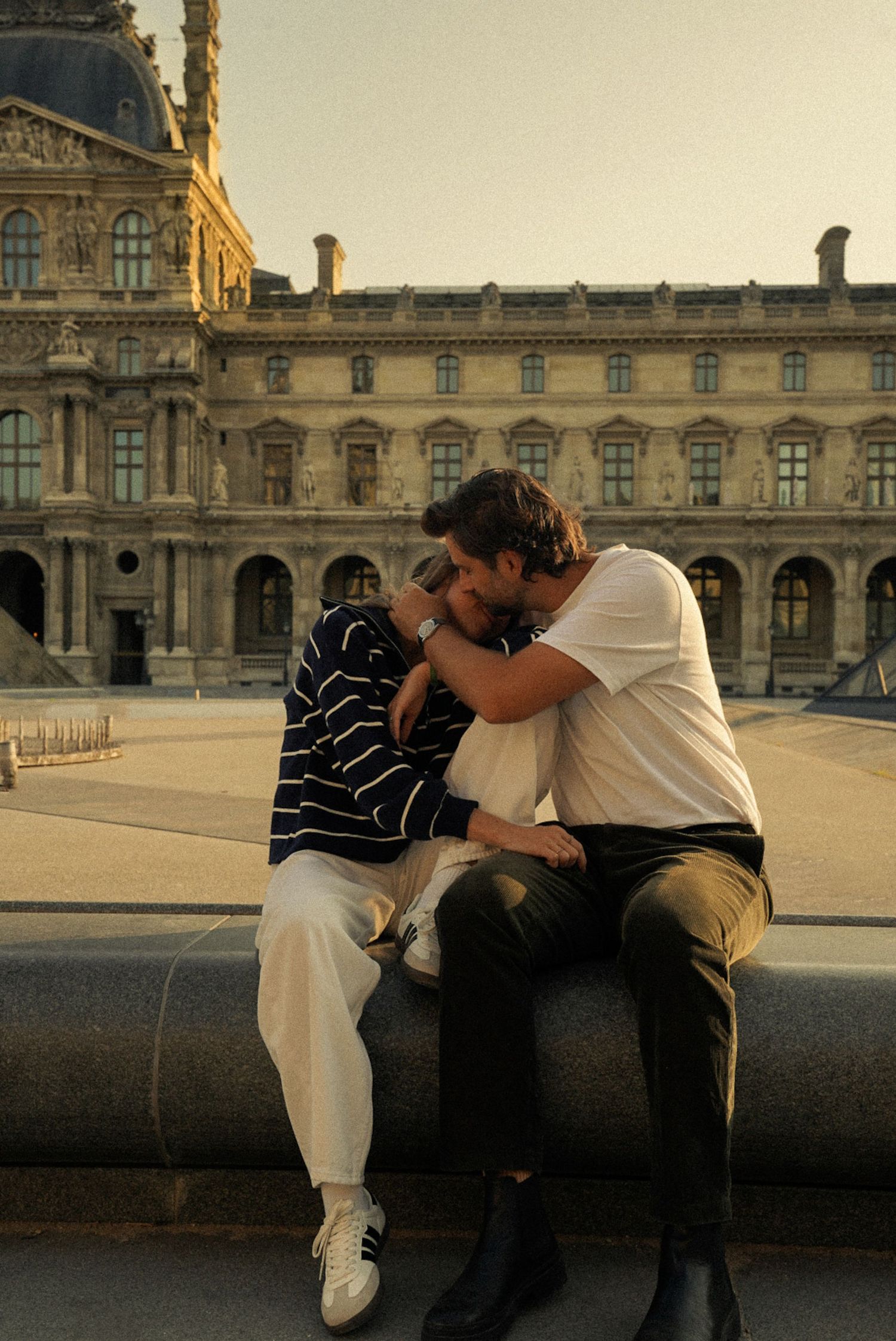 Couple sharing a romantic moment sitting together outside the Louvre Museum in Paris at golden hour.