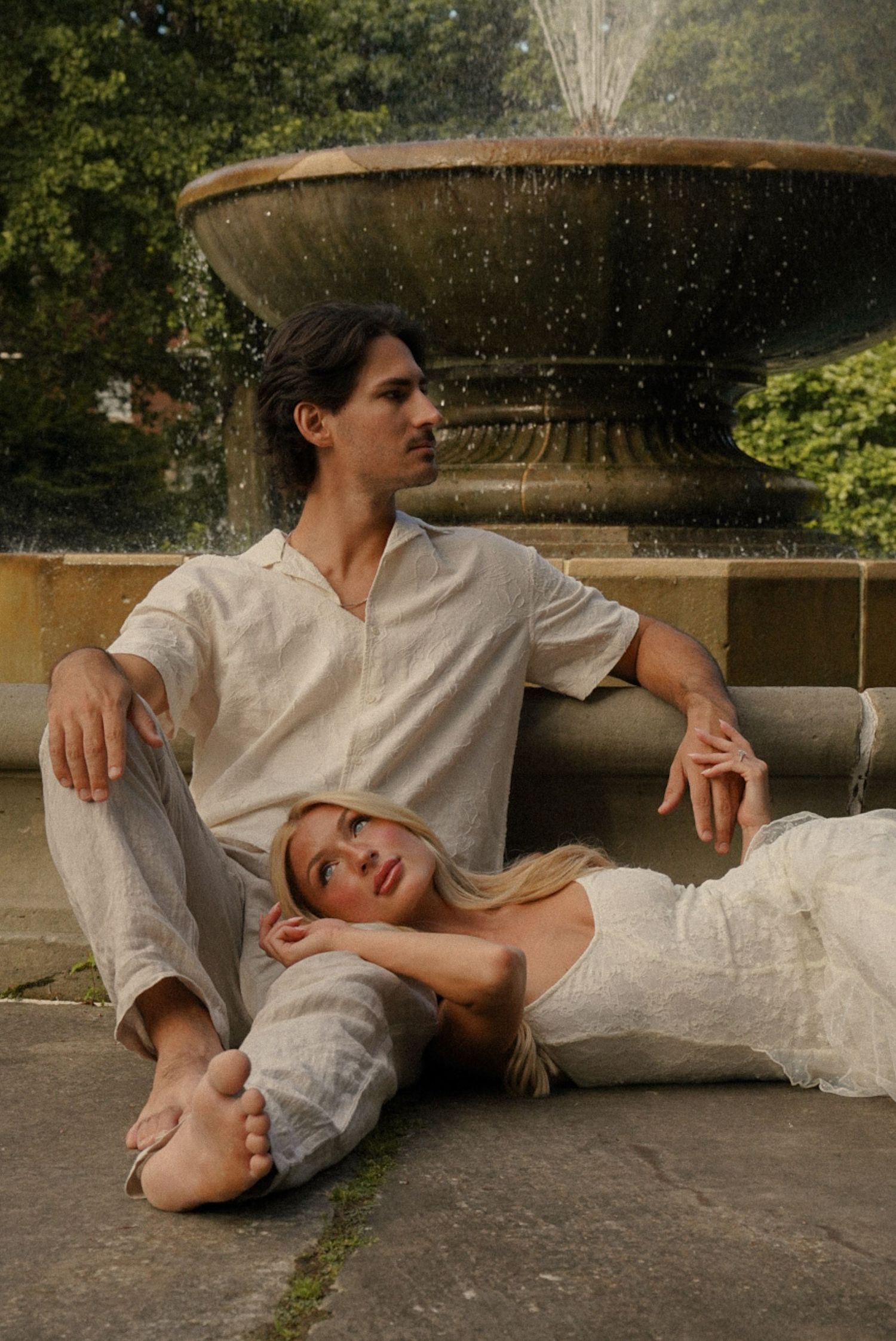 Couple in white linen outfits posing by a stone fountain in a lush green garden setting.