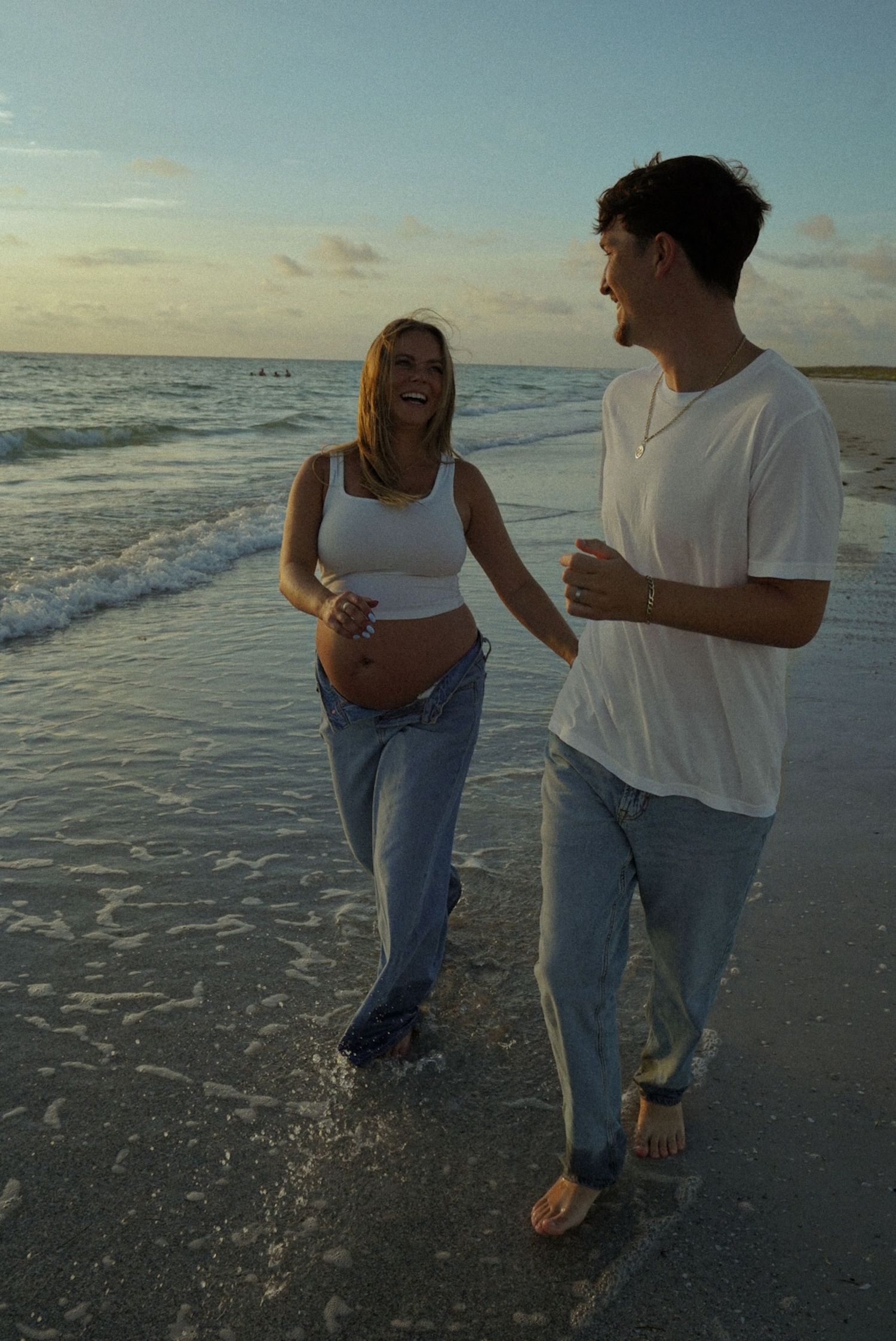 Pregnant woman and man walking barefoot on beach at sunset, laughing together in jeans and white tops.