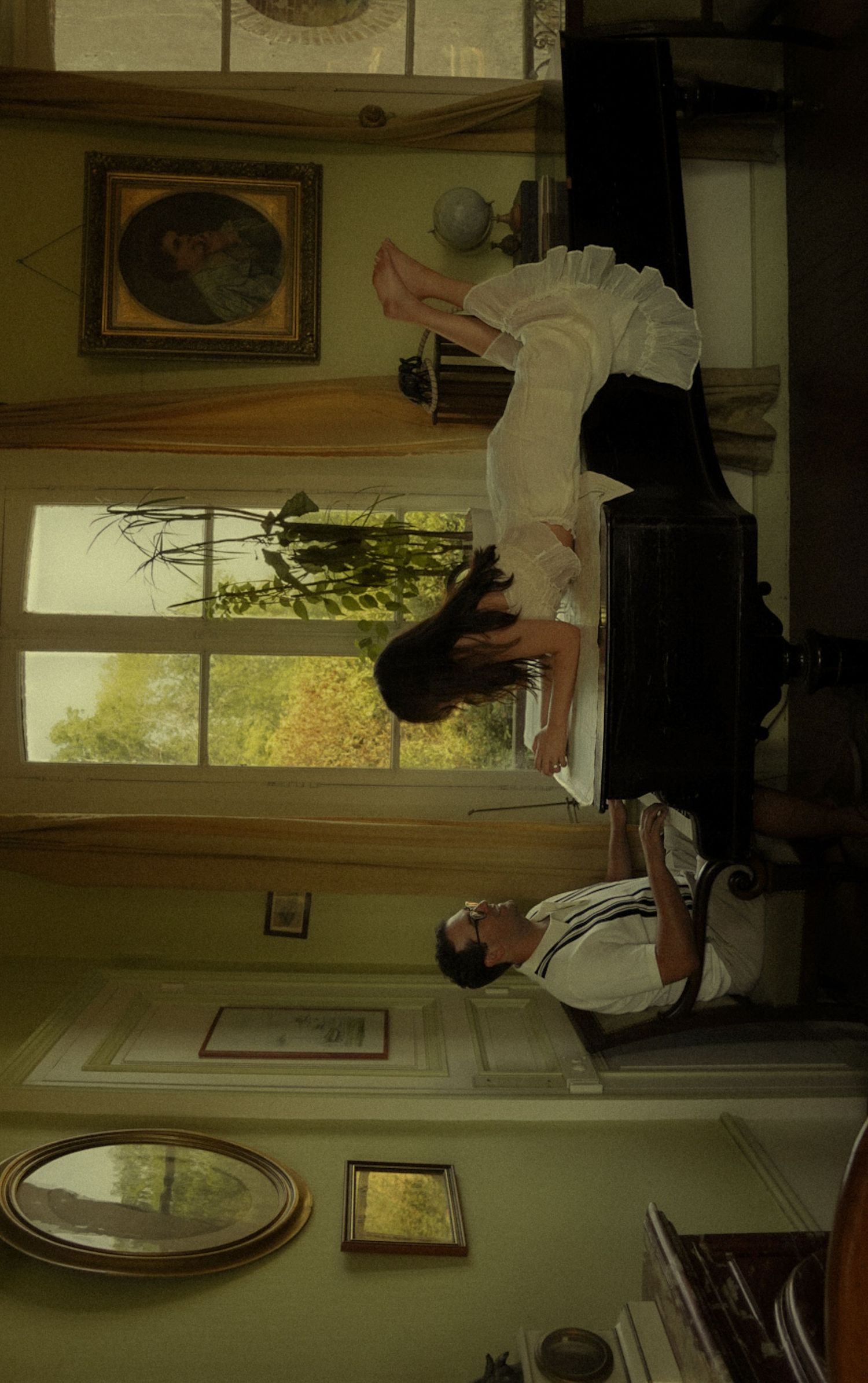 Two people in a vintage room with framed paintings, near a sunlit window showing autumn trees outside.