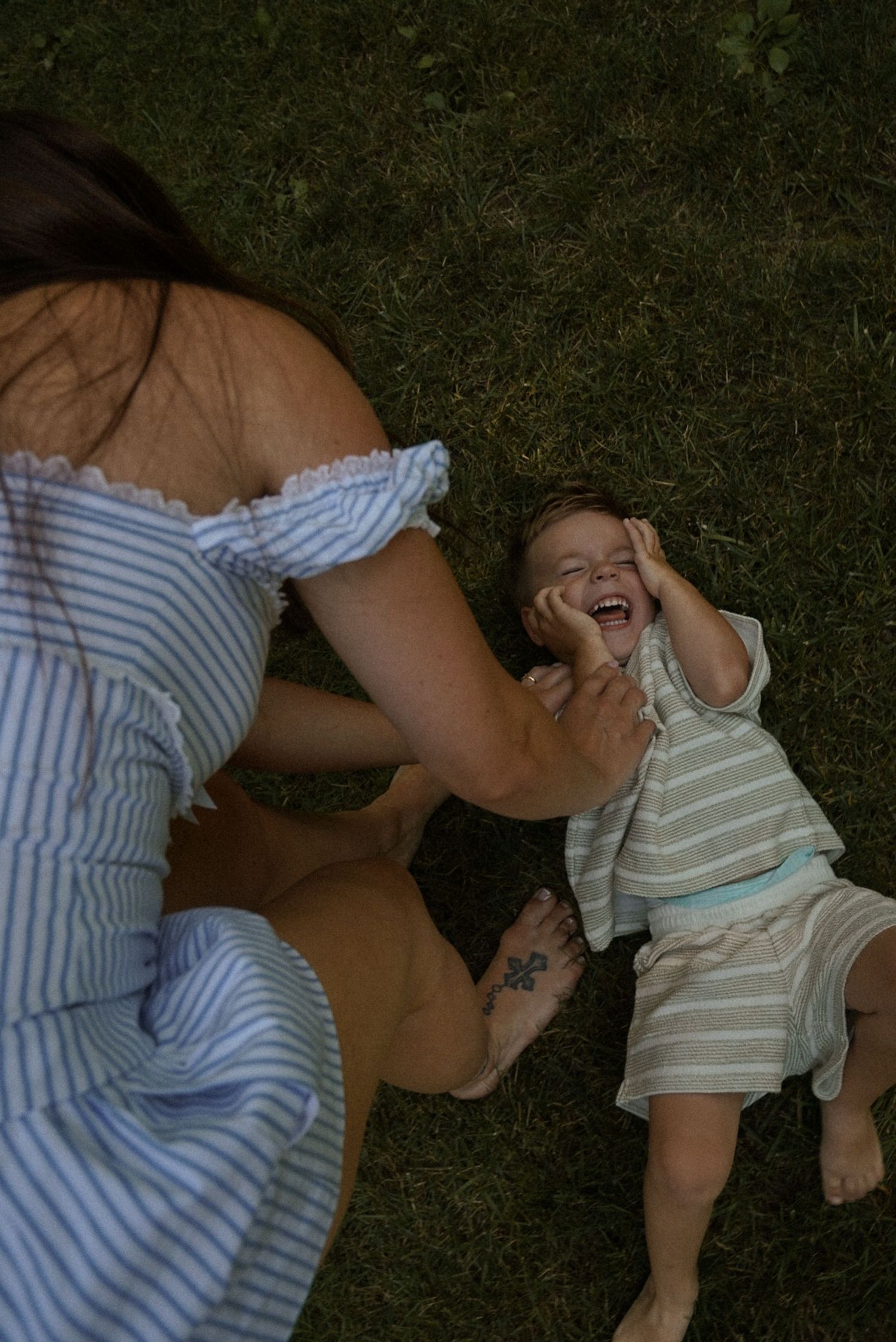 A laughing toddler lies on green grass while a woman in a blue striped off-shoulder dress plays with them outdoors.