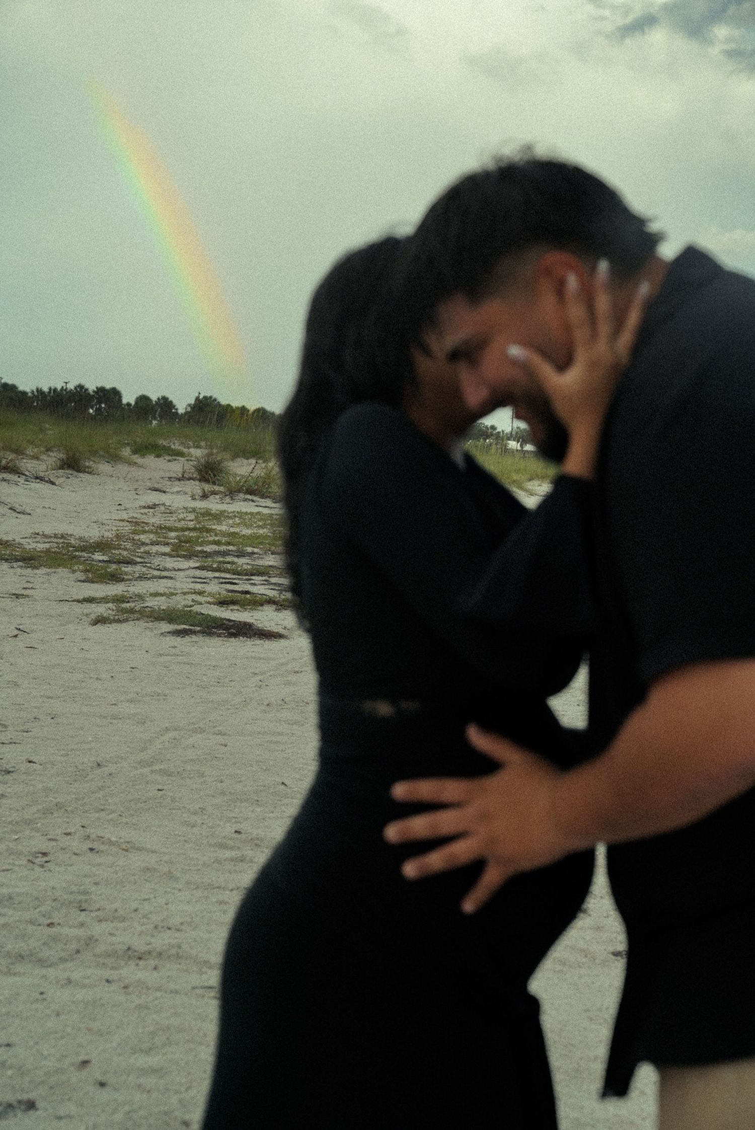 Couple in black embracing on a sandy beach with a rainbow in the cloudy sky behind them.