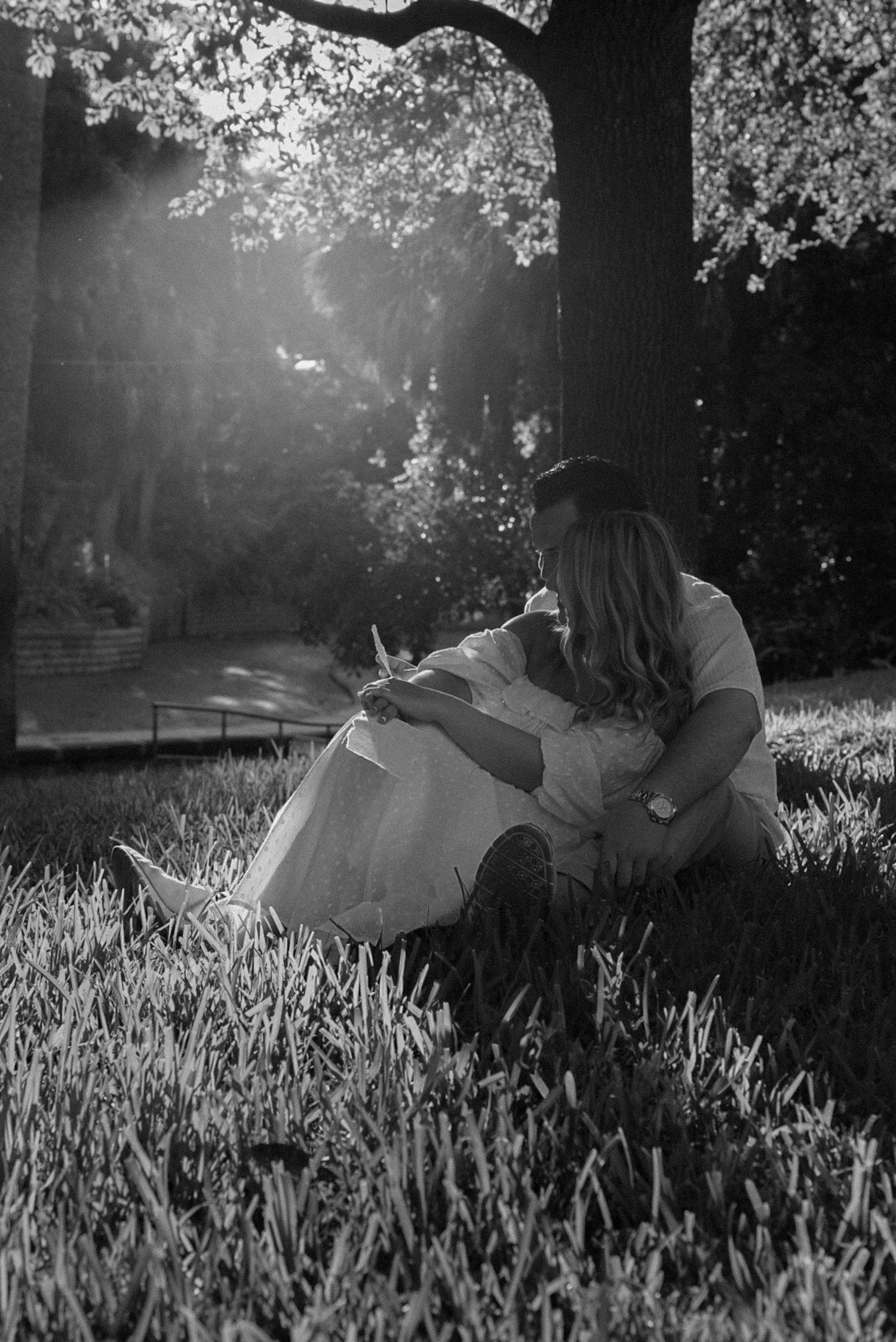Black and white photo of a couple sitting on grass under a tree, sharing a tender moment in soft sunlight.
