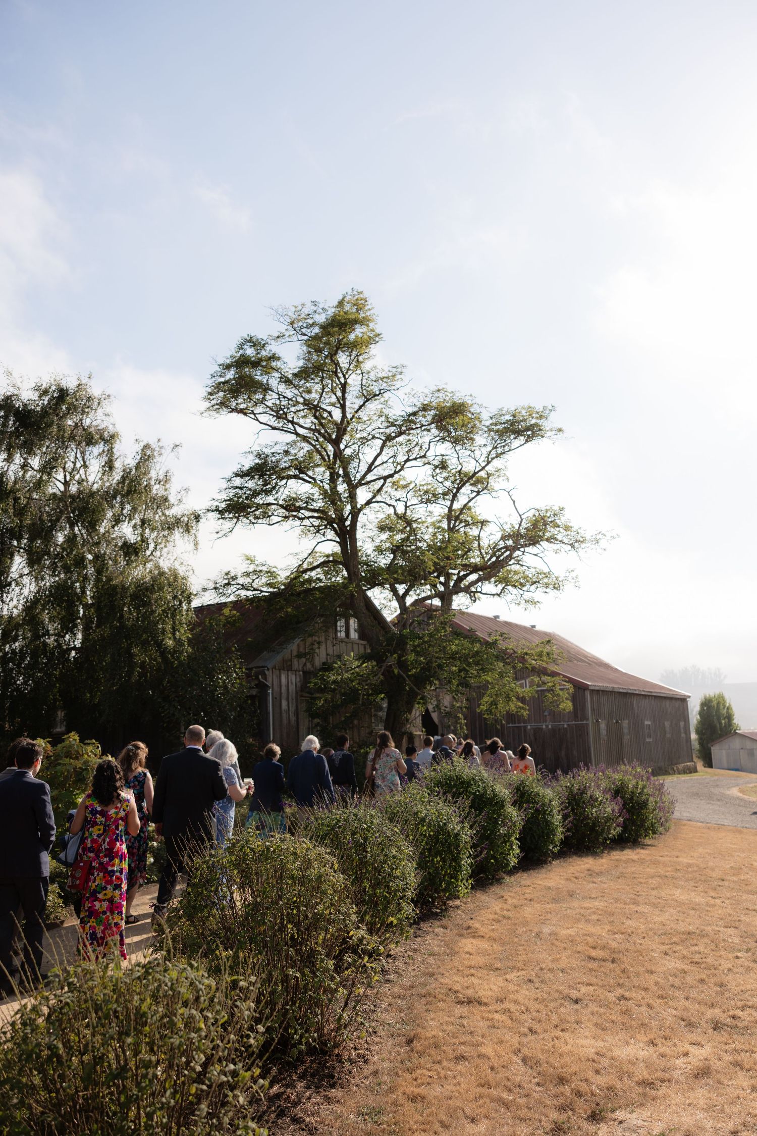 Guests walking along a garden path toward a rustic barn venue surrounded by trees on a sunny day.