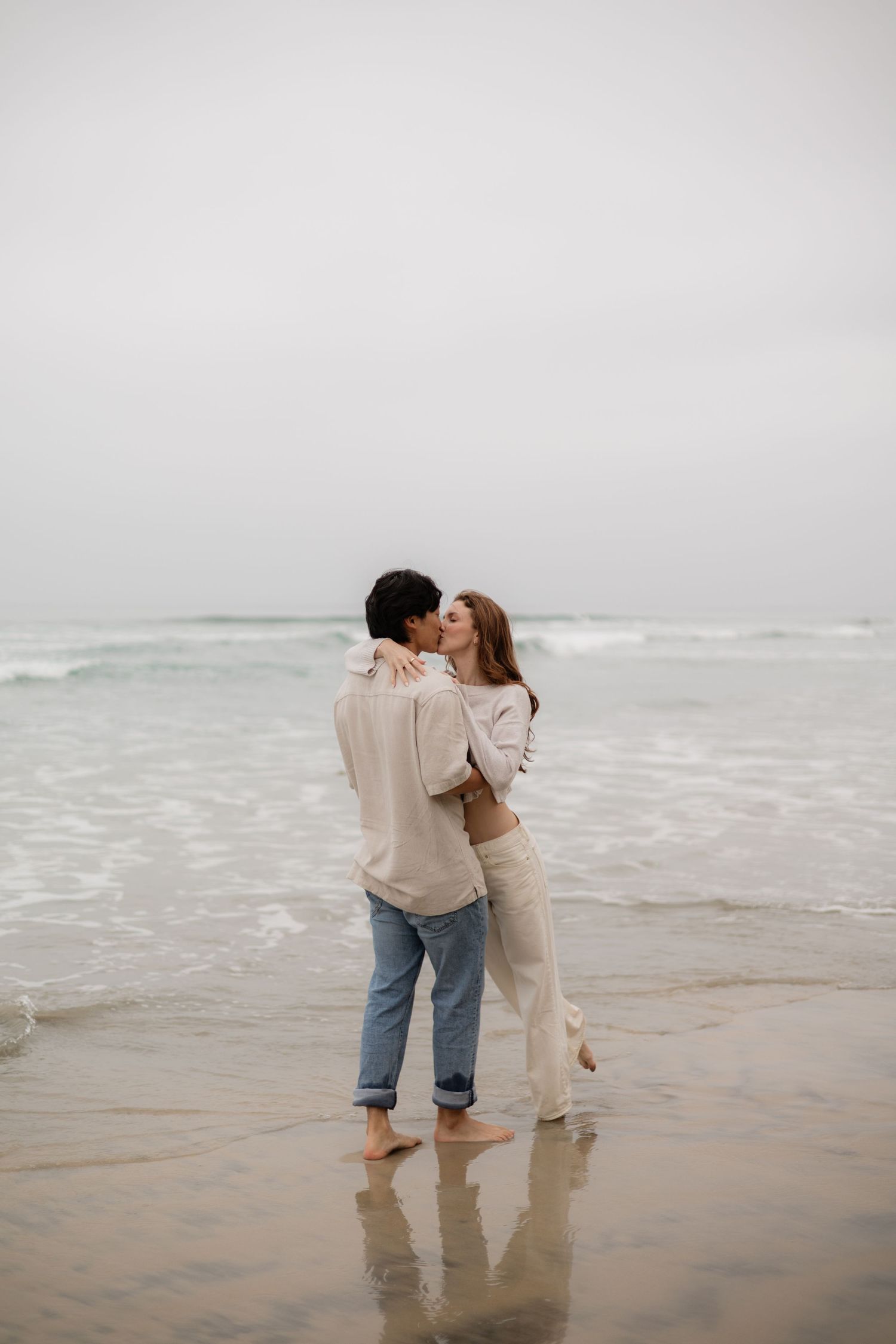 A couple shares a romantic kiss on a misty beach, standing barefoot in the shallow waves wearing casual neutral outfits.