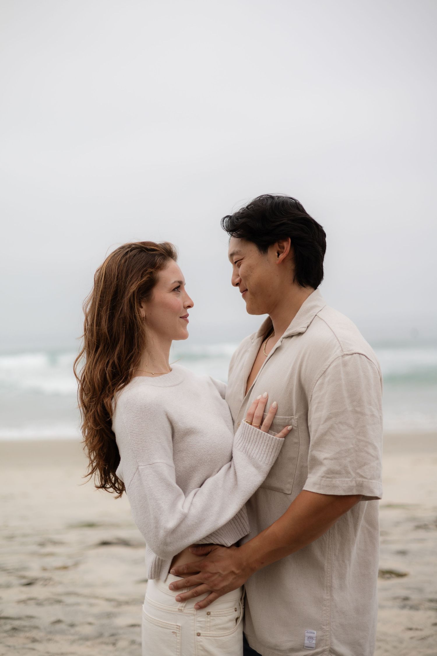 Couple facing each other in a tender embrace on a misty beach, ocean in background.