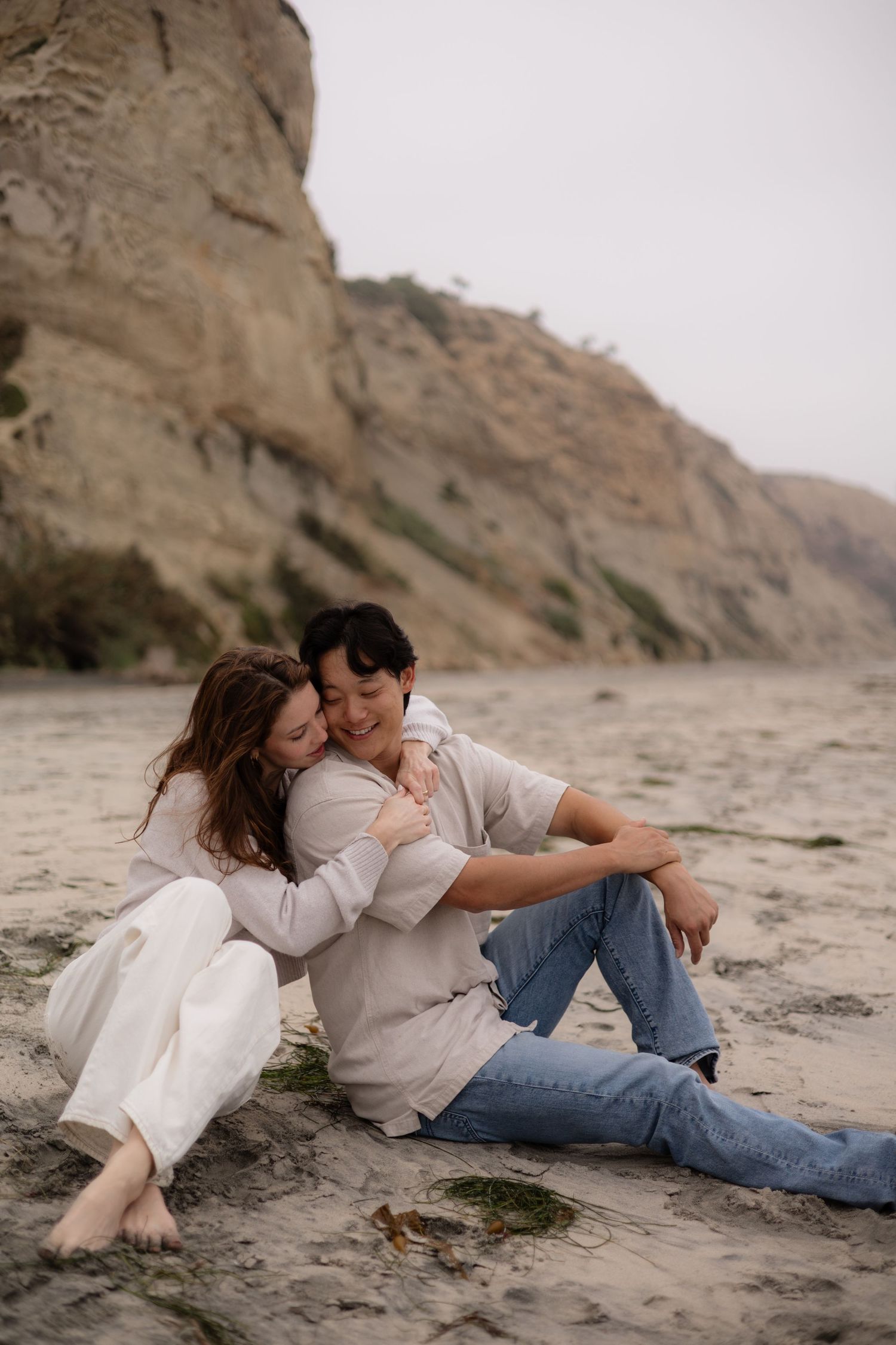 Couple sitting on sandy beach embracing, with rocky cliffs in background, wearing casual white and blue outfits.