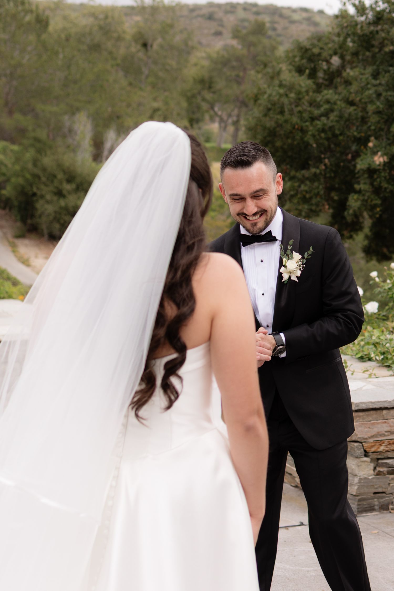 Groom in black tuxedo smiling at bride during romantic first look outdoors surrounded by lush greenery.