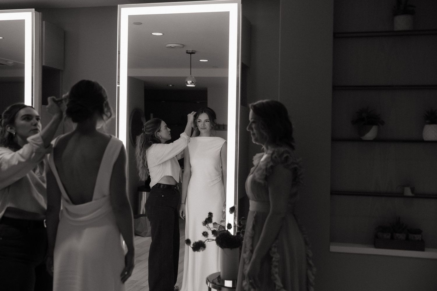 Bride getting ready with bridesmaids helping style her hair in front of a lighted mirror in black and white.