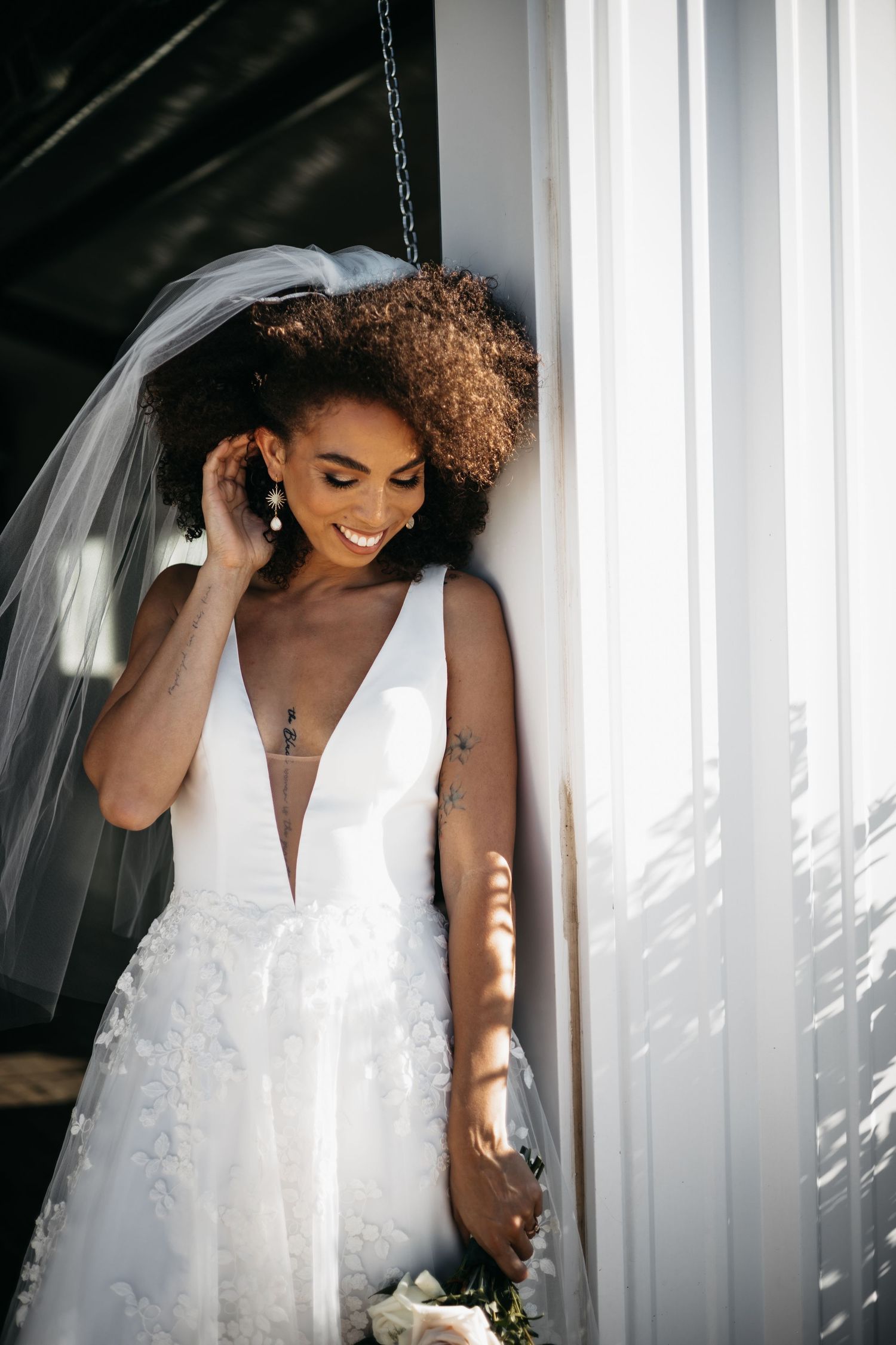 A radiant Black bride in a deep V-neck lace wedding gown and veil smiles softly in natural light.