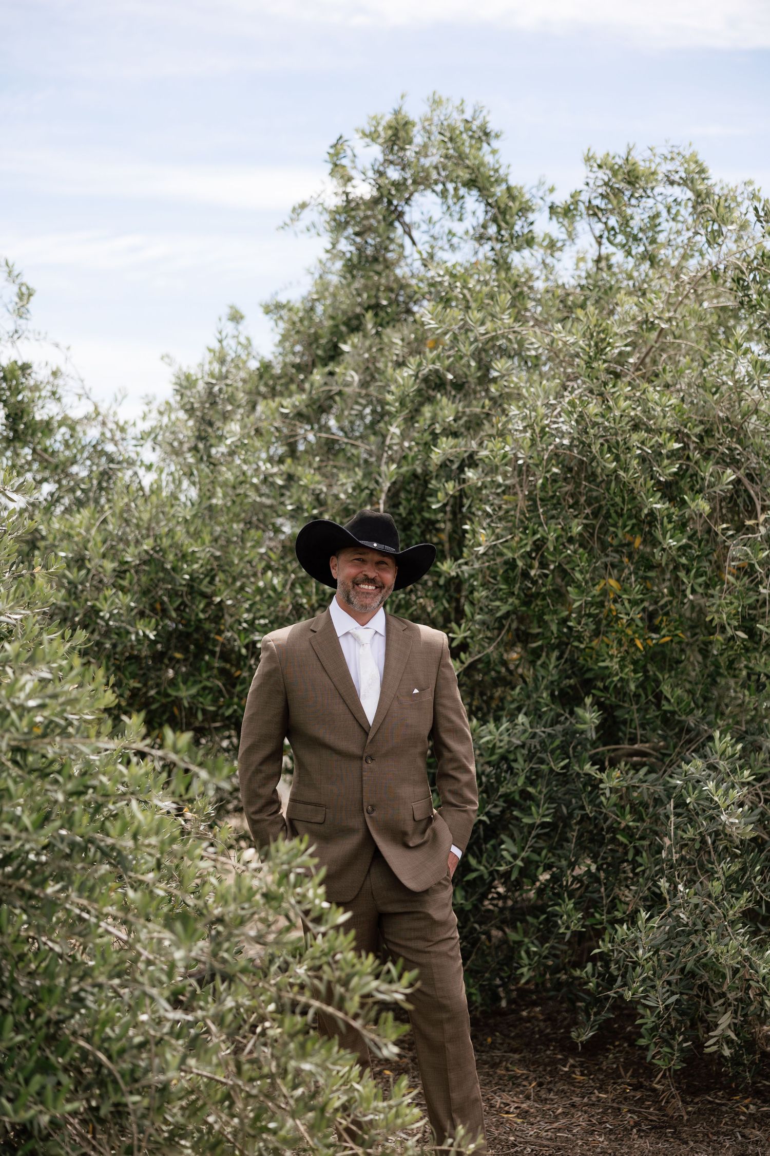 Smiling groom in brown suit and black cowboy hat posing among lush green olive trees outdoors.