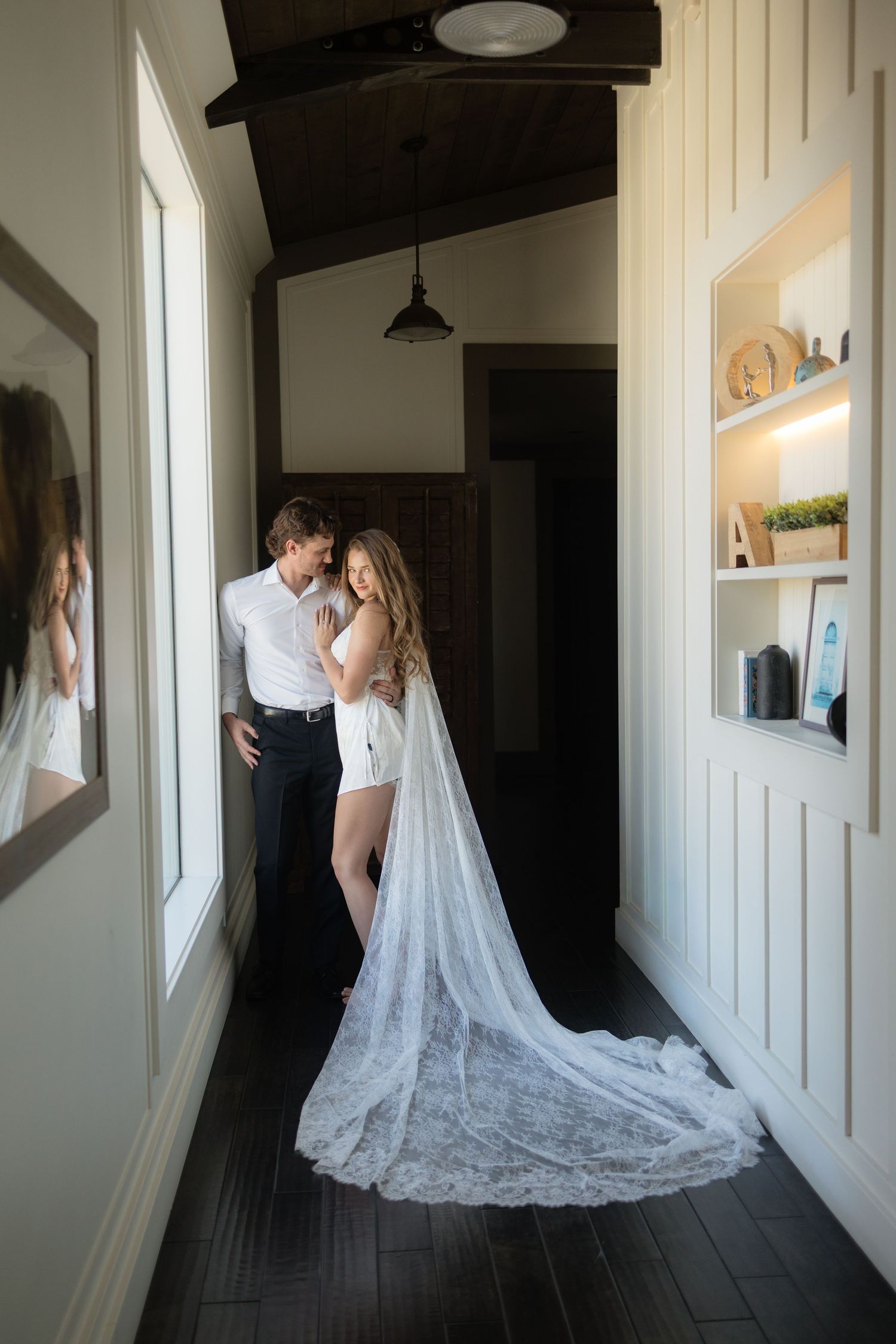 Bride in white lace veil and groom embrace tenderly in a elegant hallway with dark hardwood floors.
