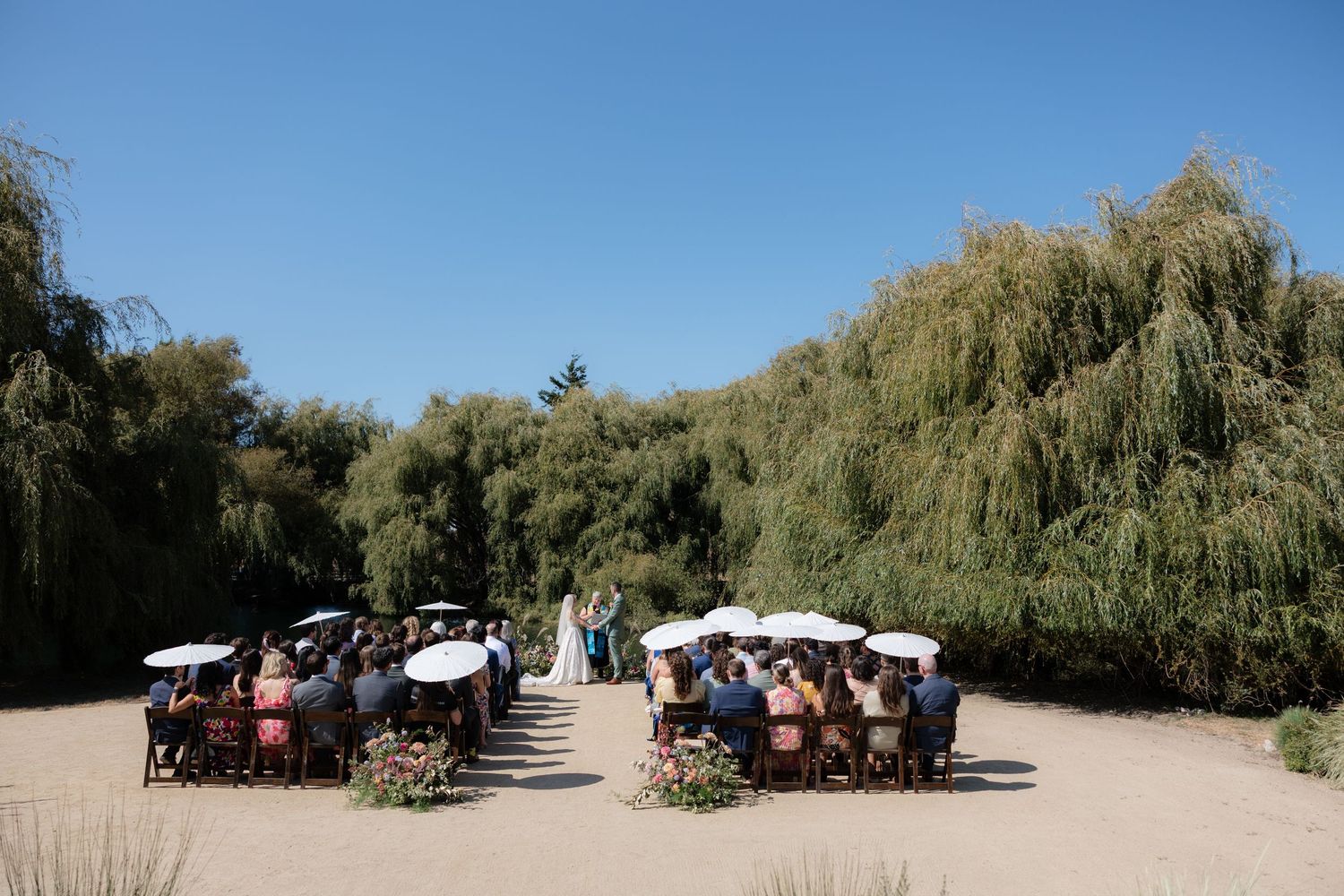 Outdoor wedding ceremony with guests seated under white umbrellas surrounded by willow trees on a sunny day.