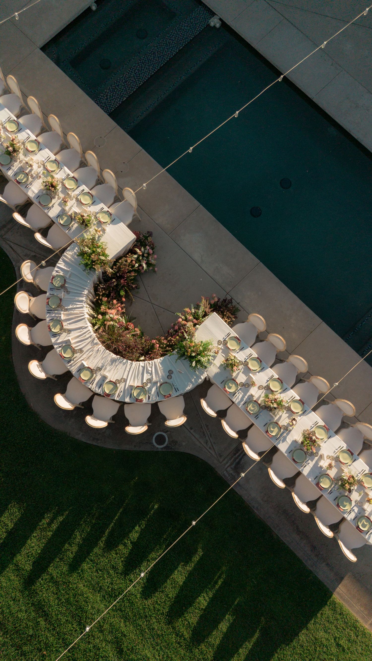 Aerial view of an elegant U-shaped dining table setup with white chairs and floral centerpieces beside a pool.