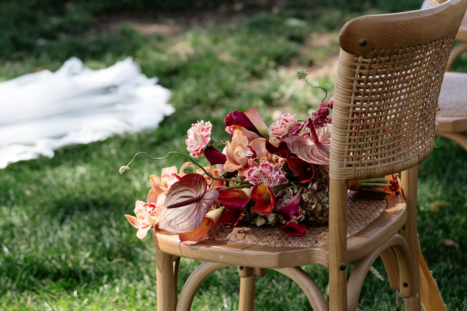 Colorful bridal bouquet with red, pink, and orange flowers resting on a rustic rattan chair on green grass.