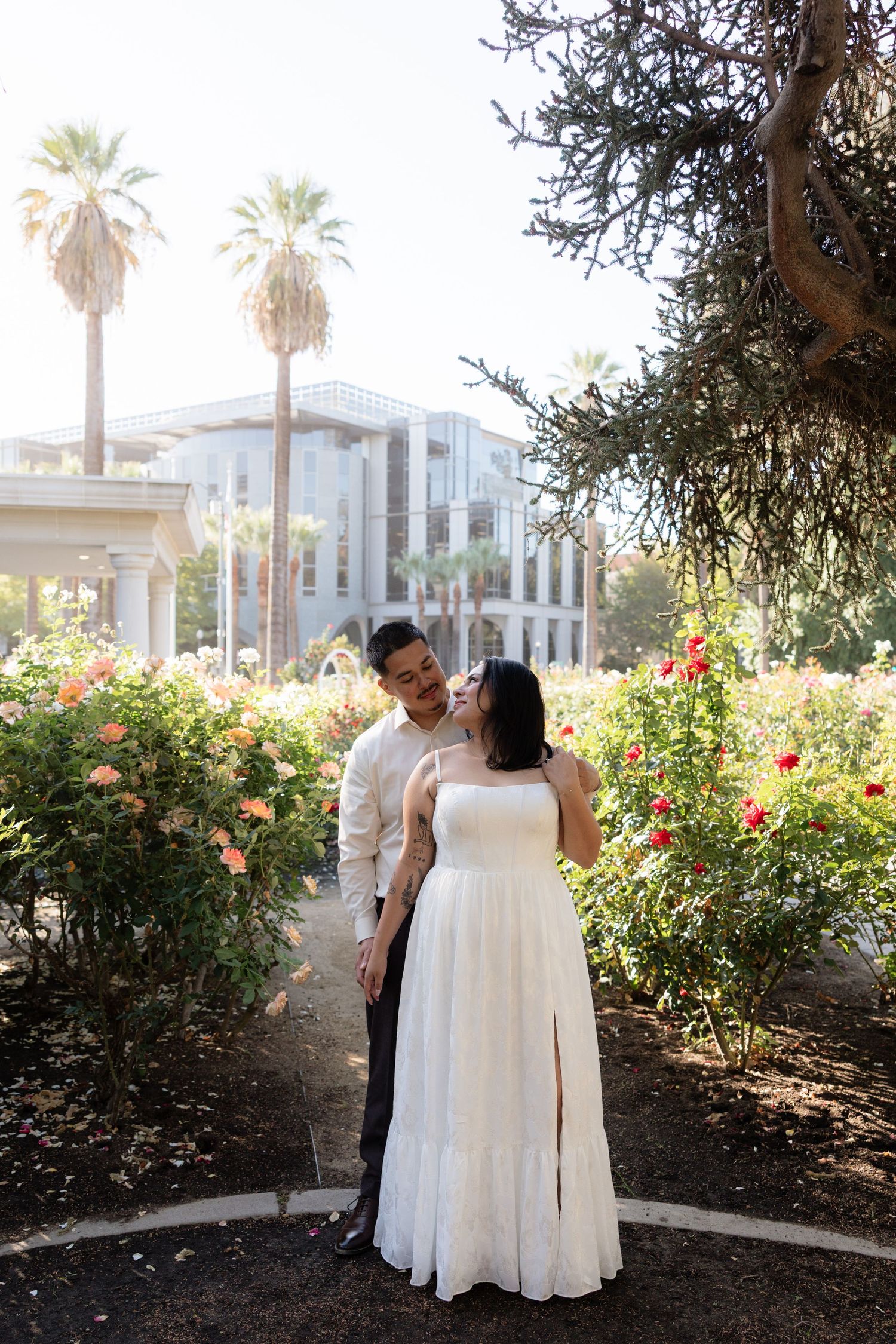 Couple in wedding attire embracing in a sunlit rose garden with palm trees and a modern building backdrop.