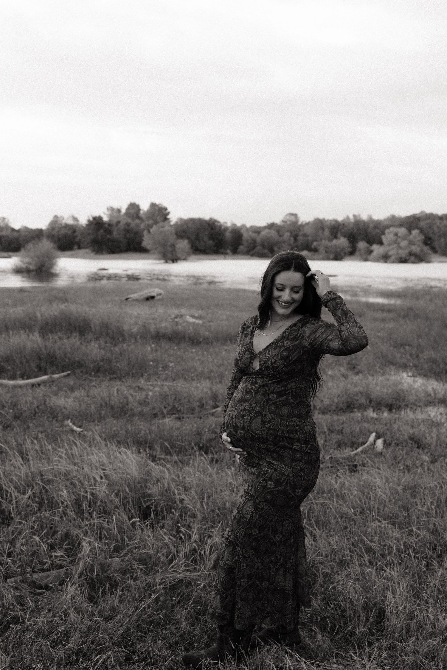 Black and white maternity photo of pregnant woman in lace gown standing in a field, smiling down at her baby bump.