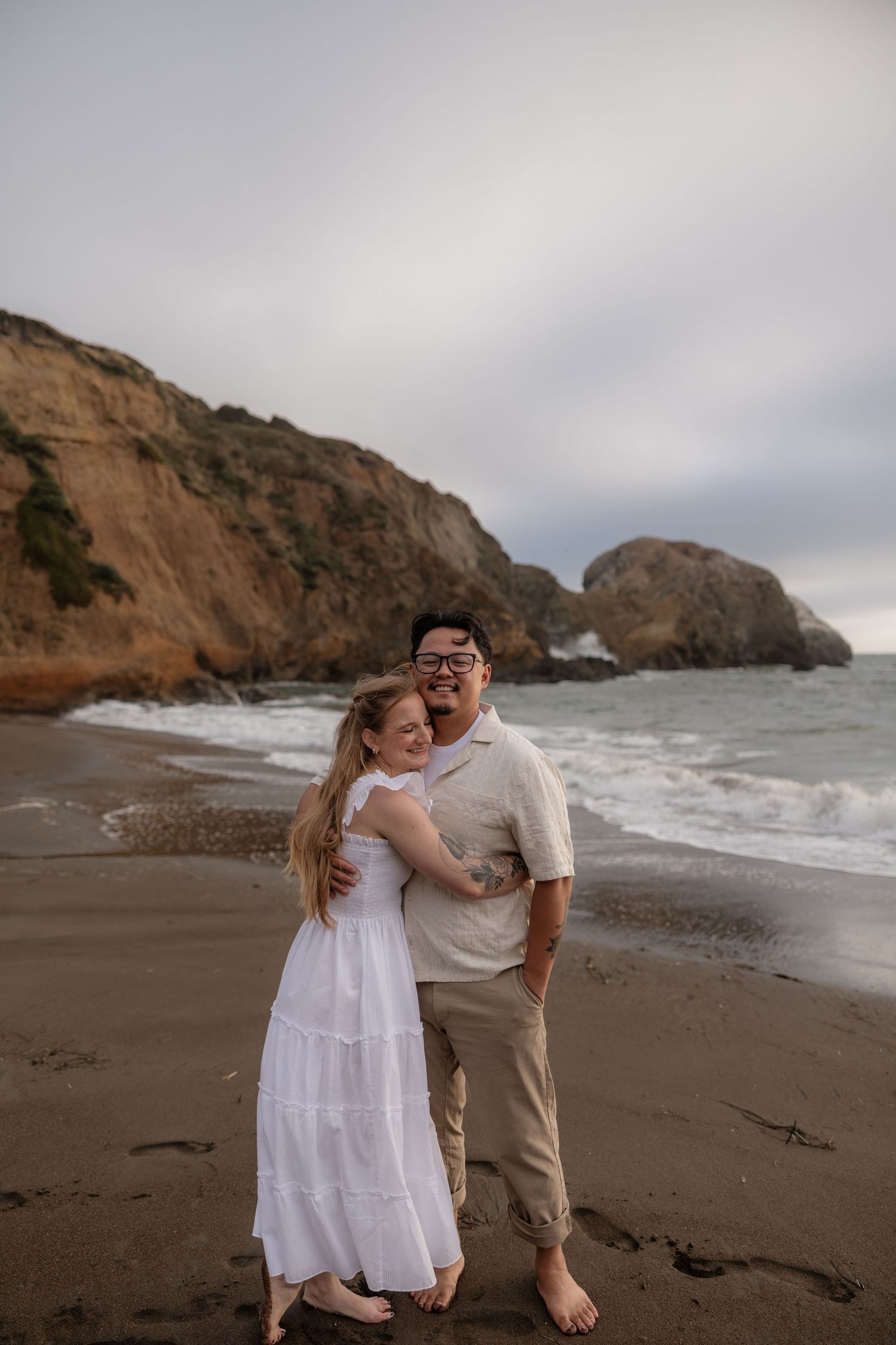 A couple embraces barefoot on a sandy beach with dramatic cliffs and ocean waves in the background.