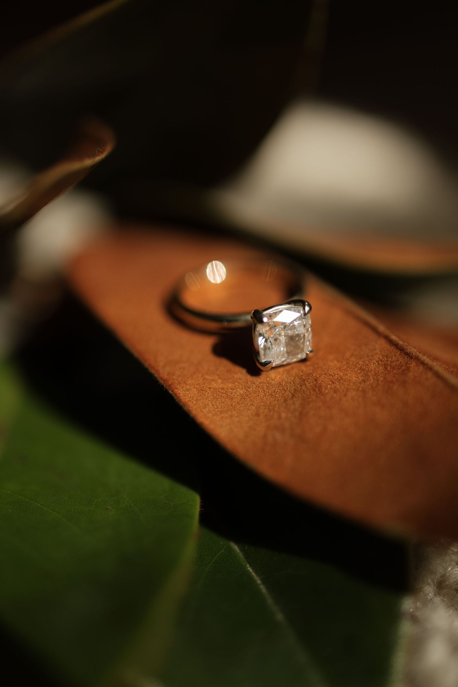 Close-up of cushion-cut diamond solitaire engagement ring resting on brown leather with green leaves.