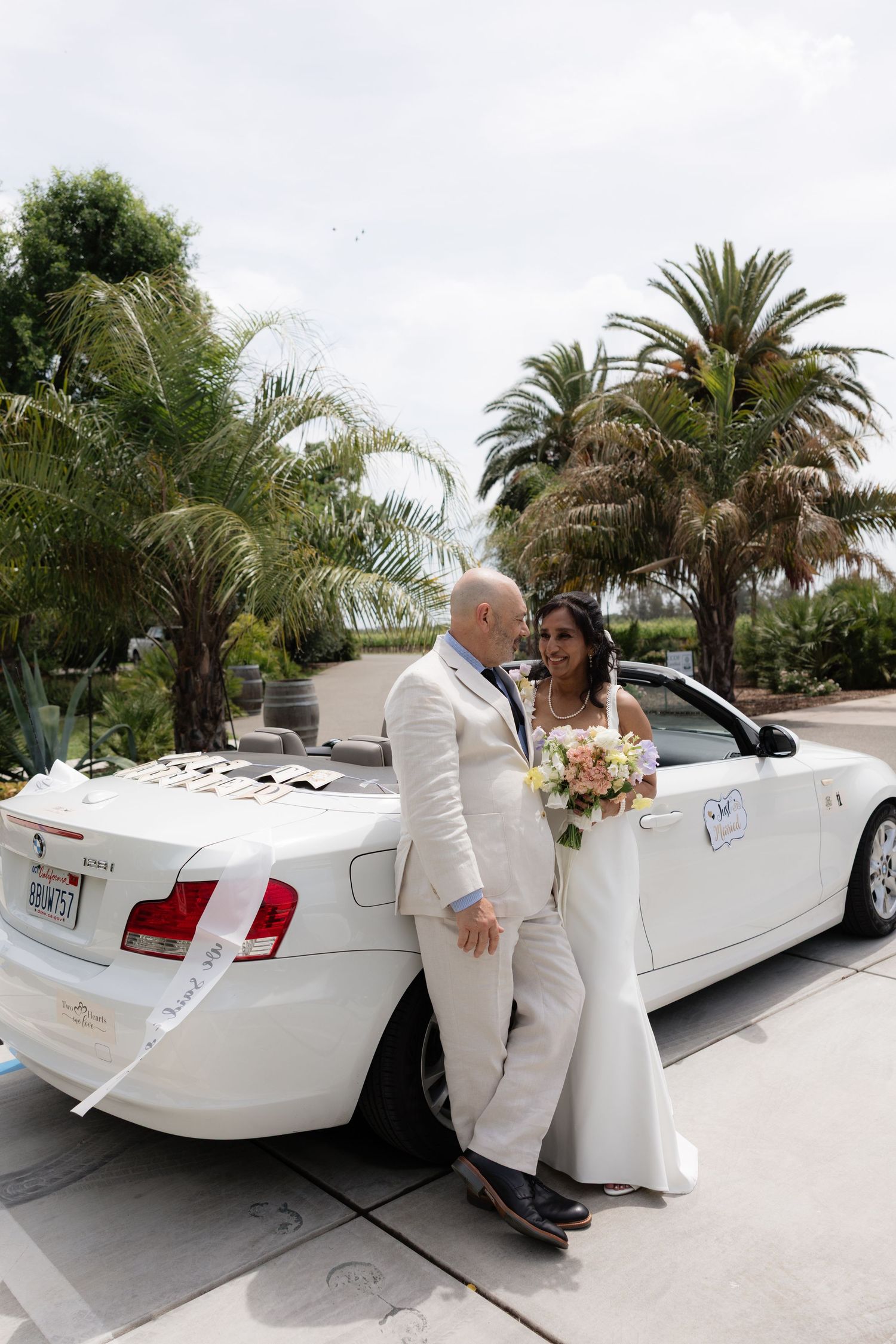 Bride and groom lean against decorated white convertible BMW getaway car surrounded by palm trees after wedding ceremony.