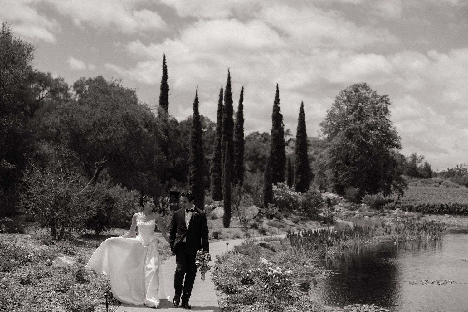 Black and white photo of bride and groom walking along a garden path beside a lake with tall cypress trees.