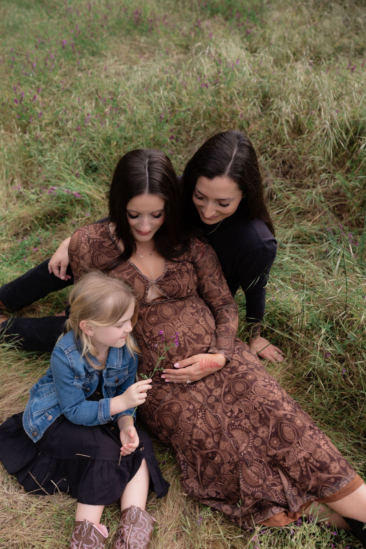 Pregnant woman in brown dress sitting in a field with her partner and young daughter.