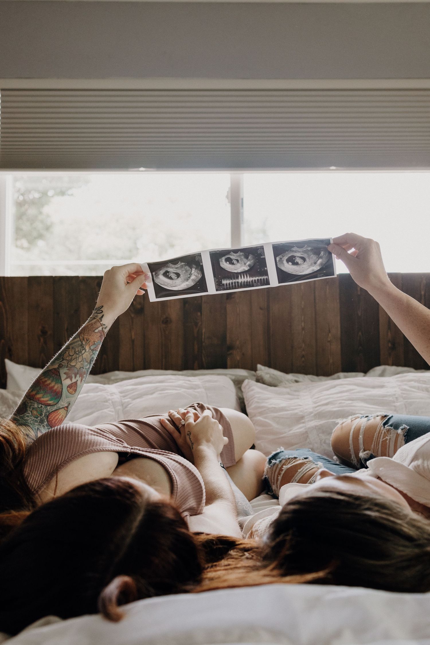 Couple lying on bed holding ultrasound photos, pregnancy announcement with tattooed partner.