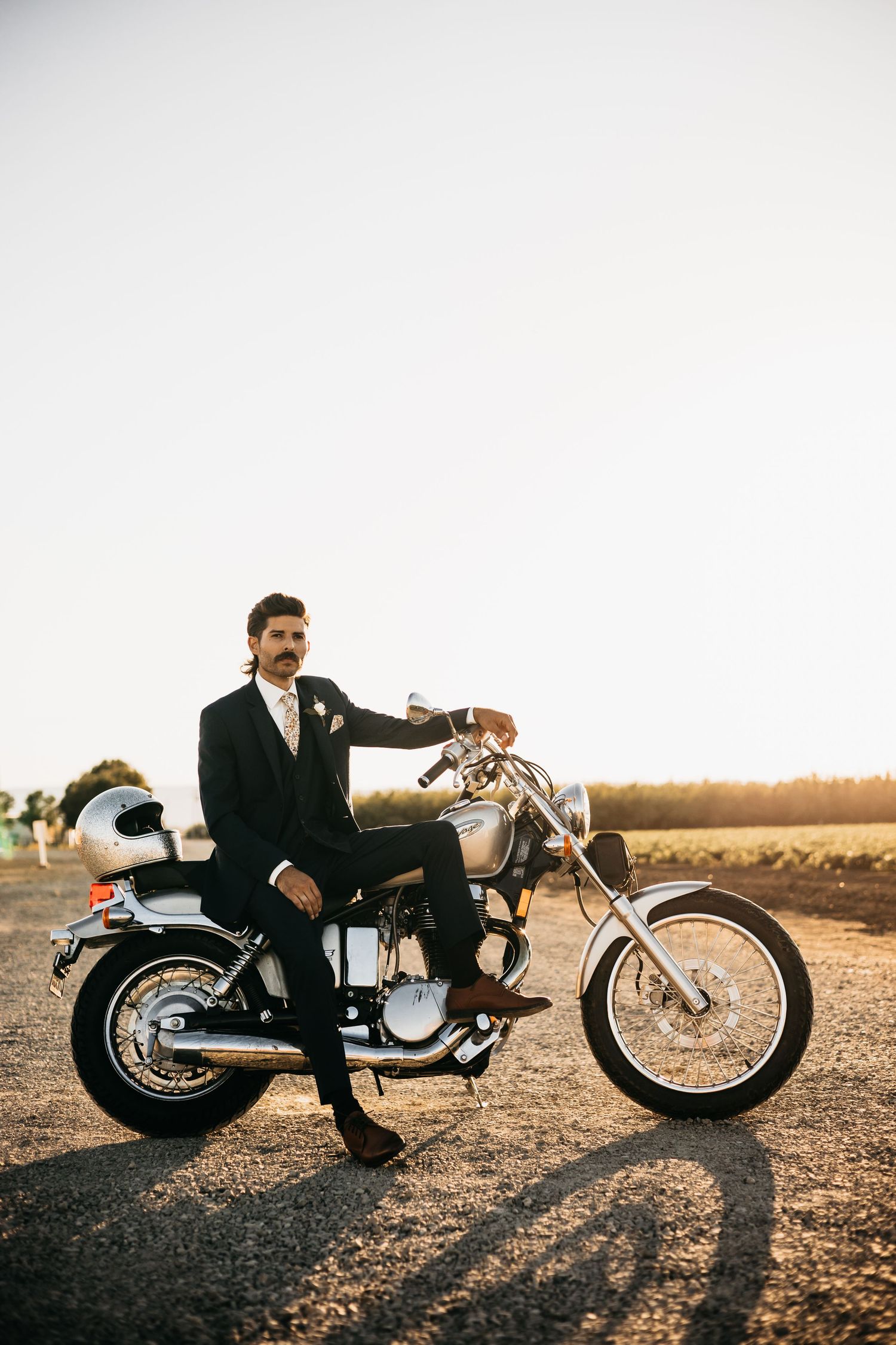 A dapper groom with a mustache sits confidently on a classic motorcycle at sunset in a black suit.