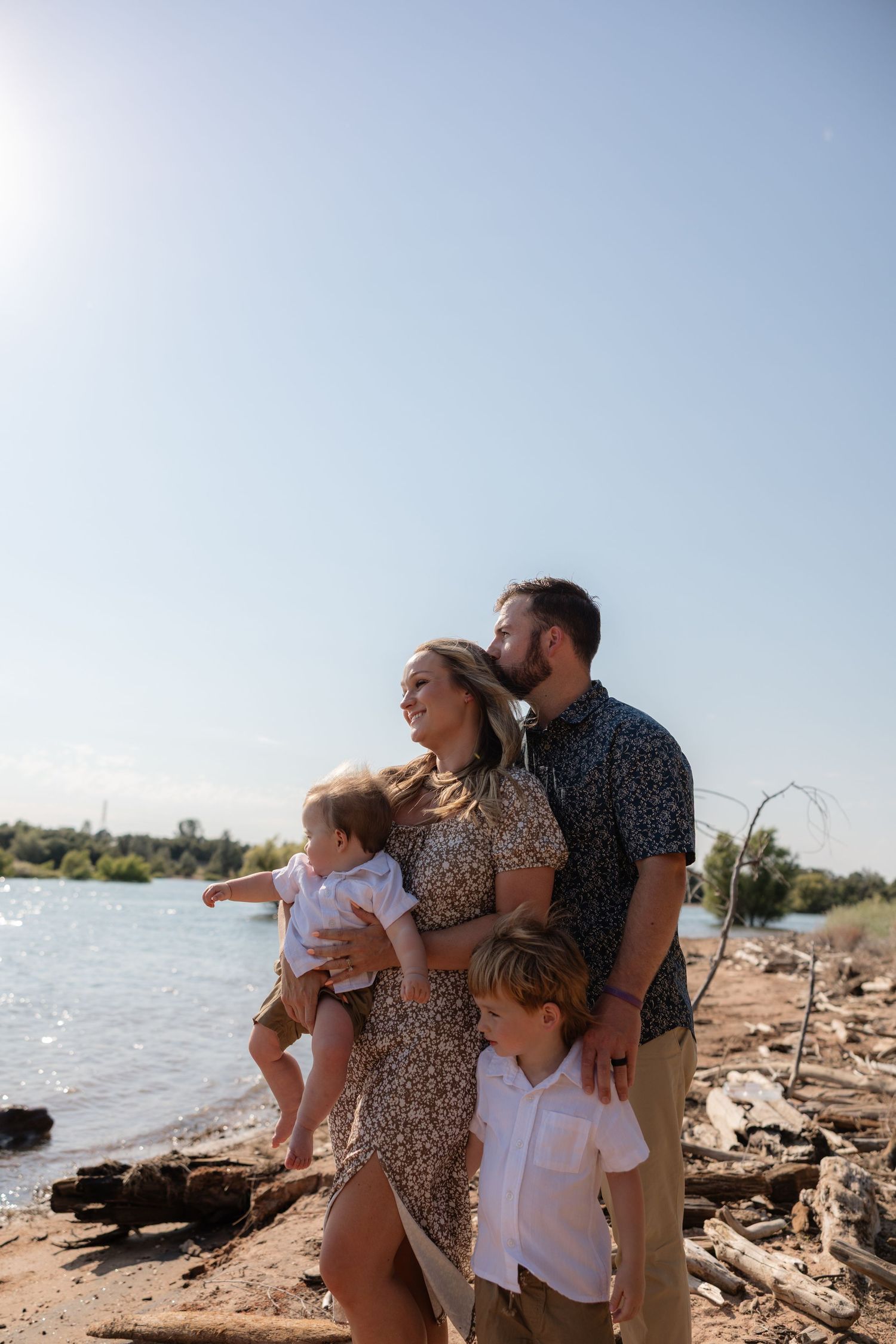 Family of four posing near a sunny lakeside, parents holding young children on rocky shore with trees in background.