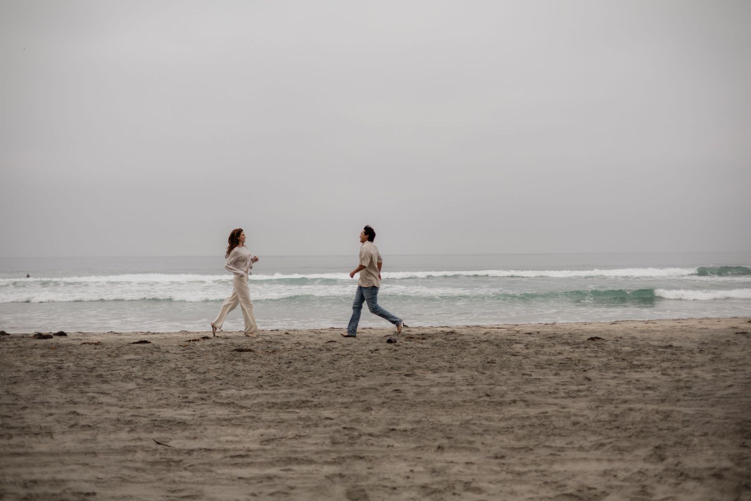 Two women walking on a sandy beach with overcast skies and ocean waves in the background.