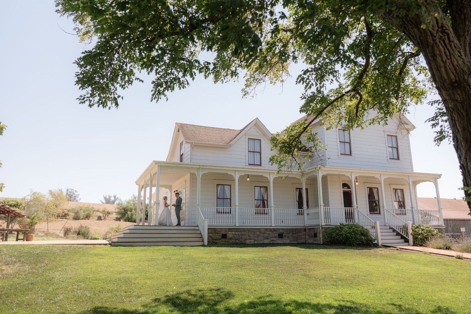 Historic white two-story Victorian farmhouse with wrap-around porch, green lawn, and large shade tree on sunny day.
