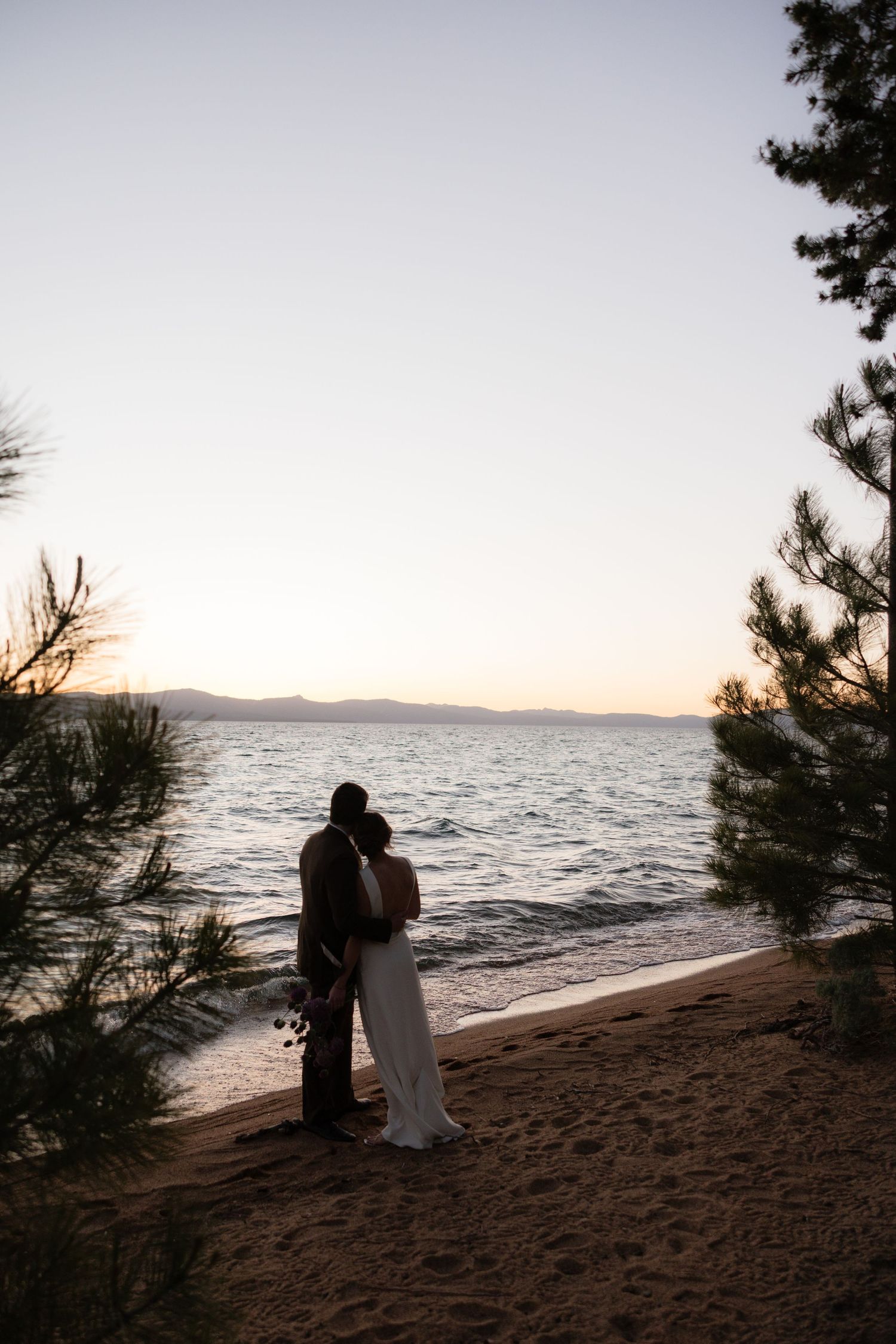 Couple silhouetted against a serene lake sunset on a sandy beach, framed by pine trees.