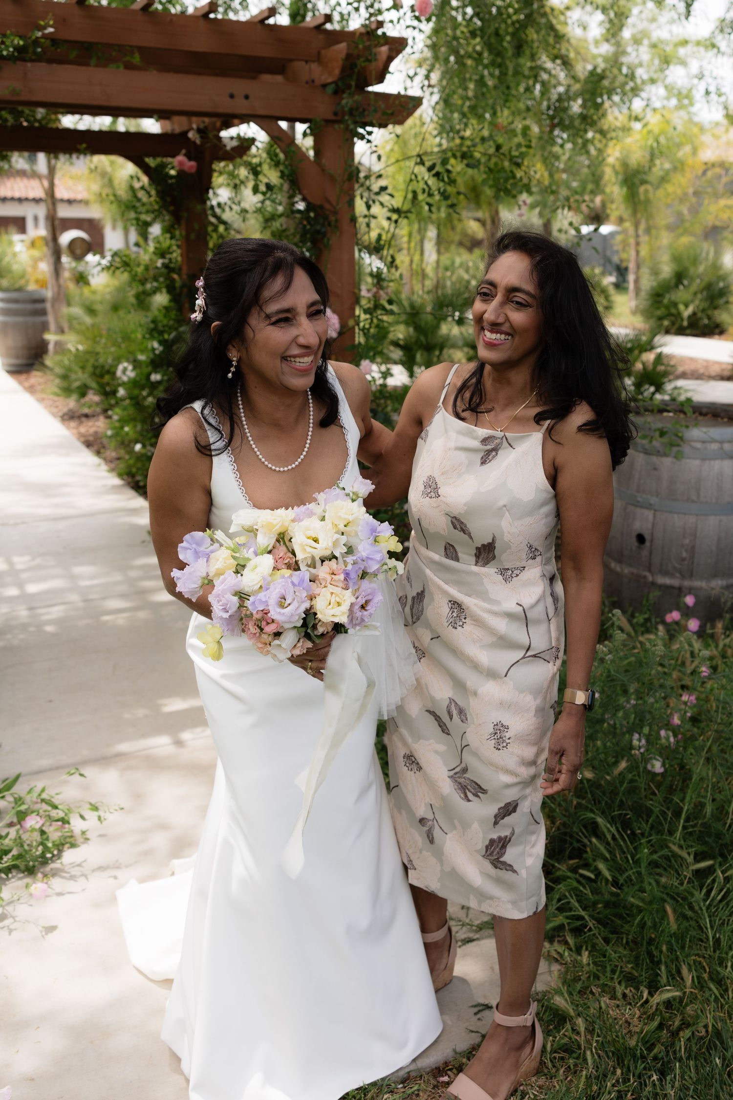 Smiling bride holds pastel bouquet while walking with female guest under wooden pergola in garden.