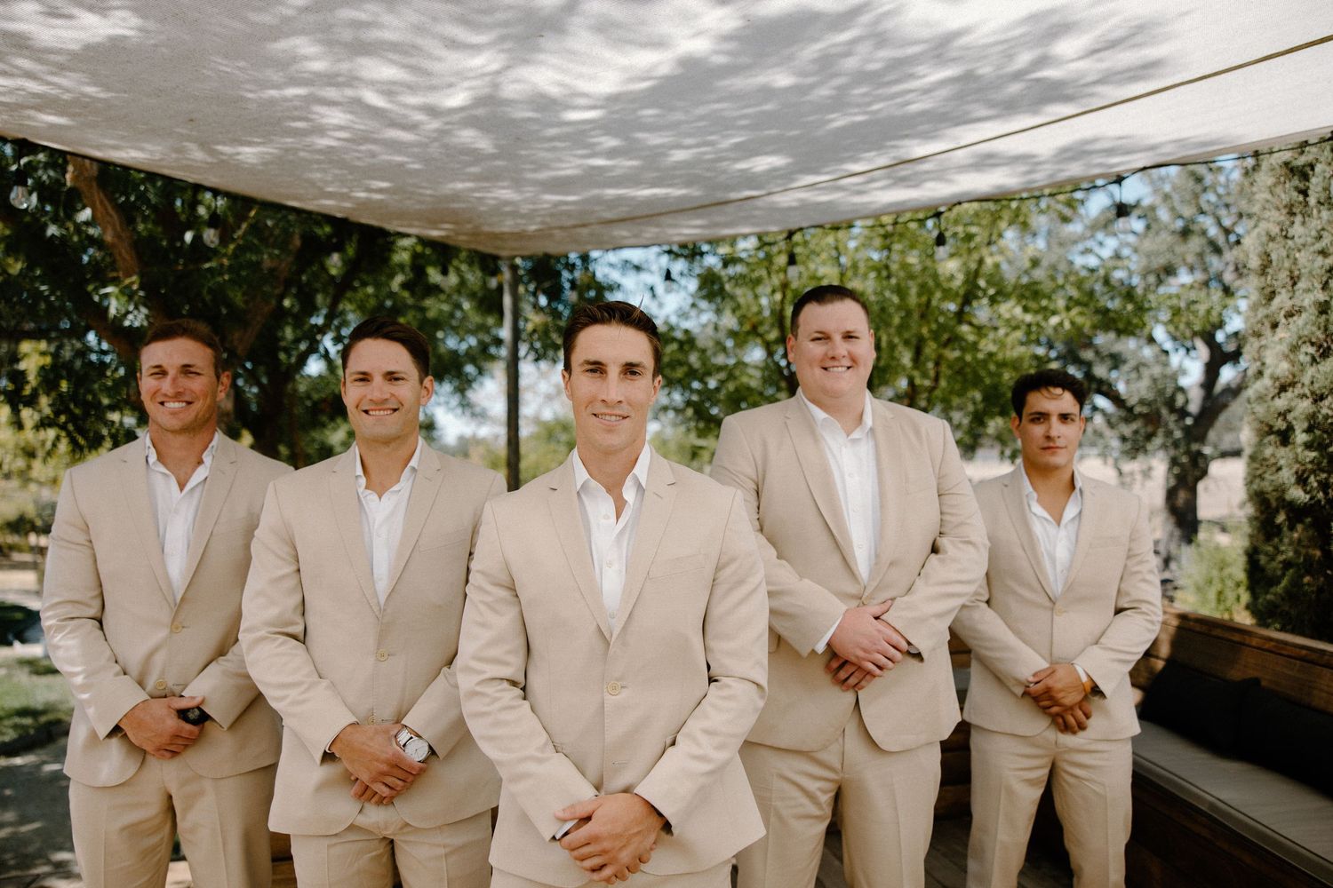 Five groomsmen in matching tan suits posing confidently under a pergola at an outdoor wedding.