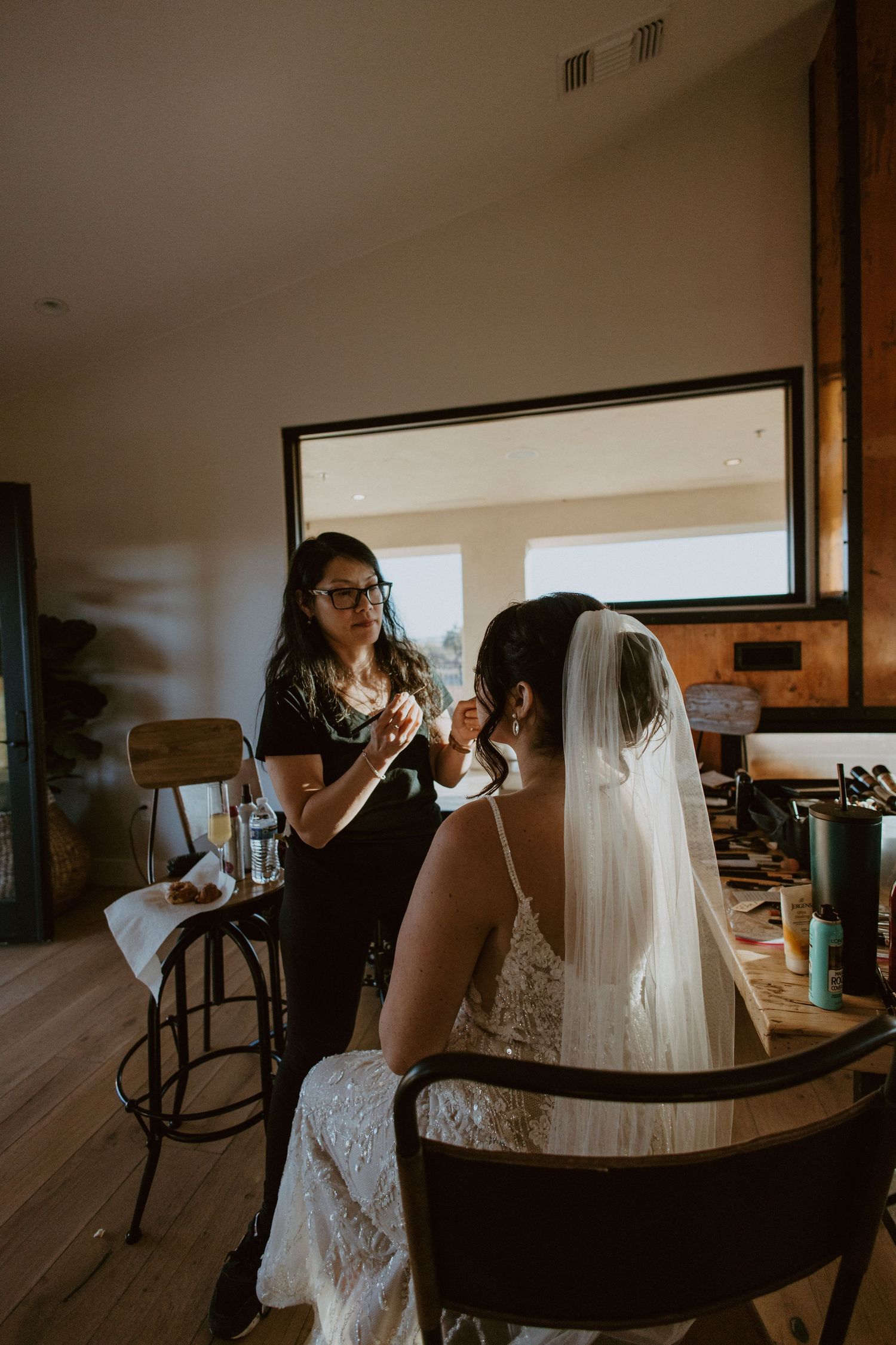Makeup artist applying finishing touches to bride wearing lace gown and veil before ceremony.
