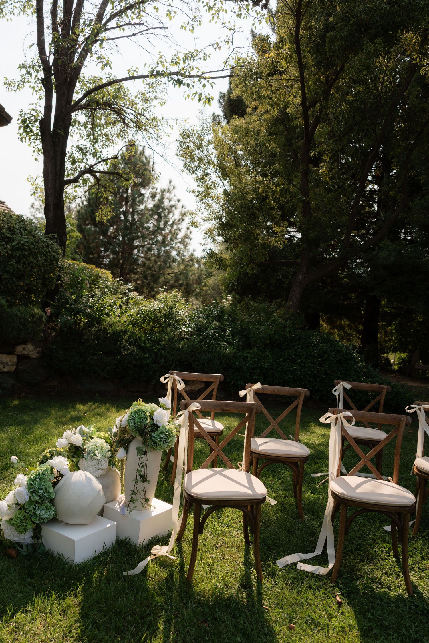 Outdoor wedding ceremony setup with wooden cross-back chairs on green lawn surrounded by lush trees and white floral arrangements.