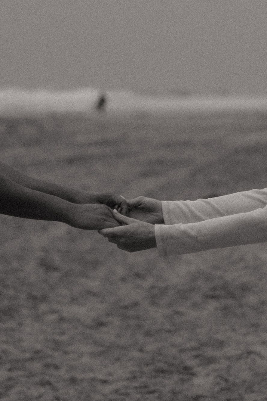 Black and white photo of two people gently holding hands on a sandy beach with ocean waves in the background.