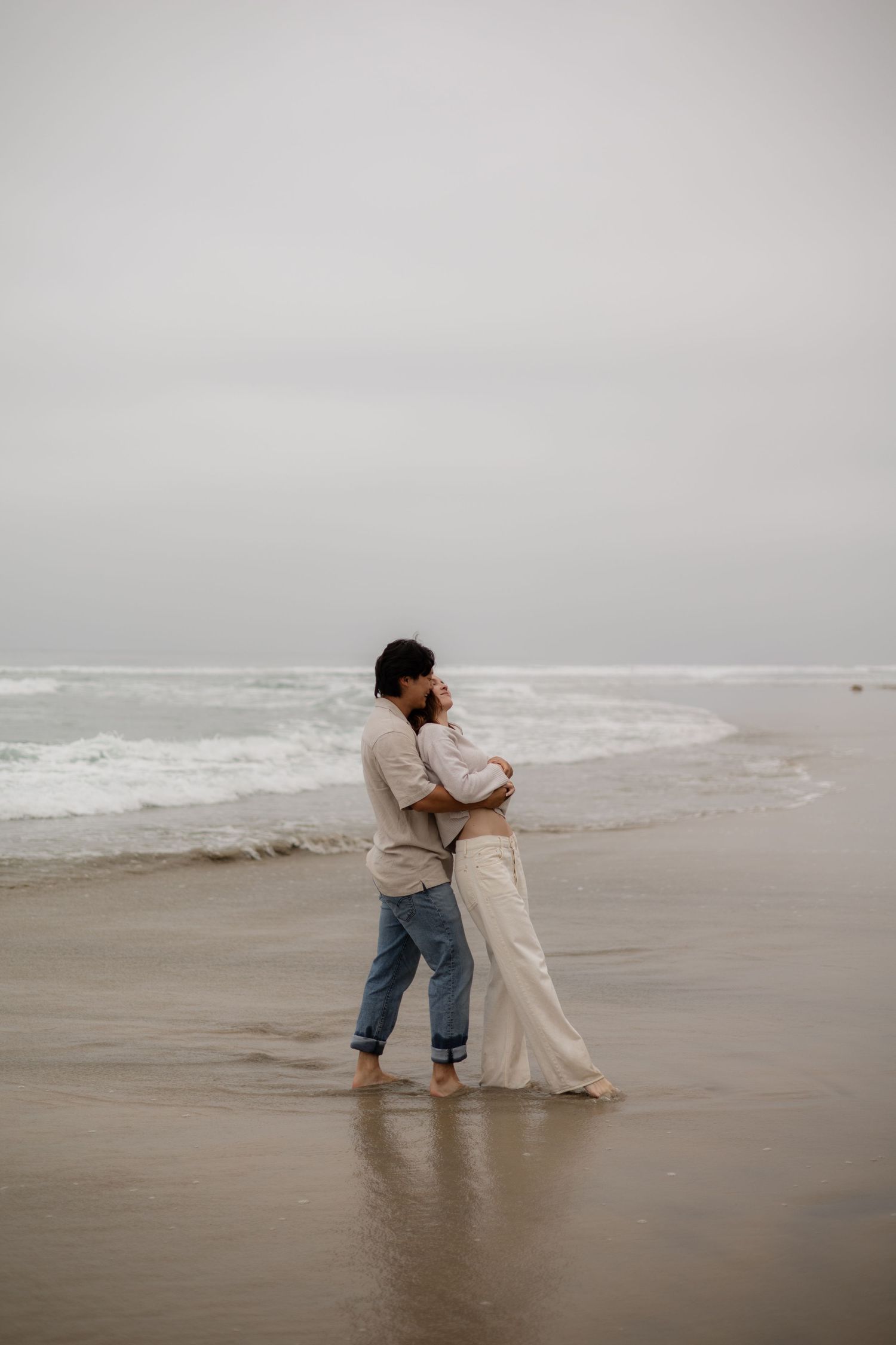 Couple embracing barefoot on a misty beach, wearing casual neutral tones, with ocean waves in the background.