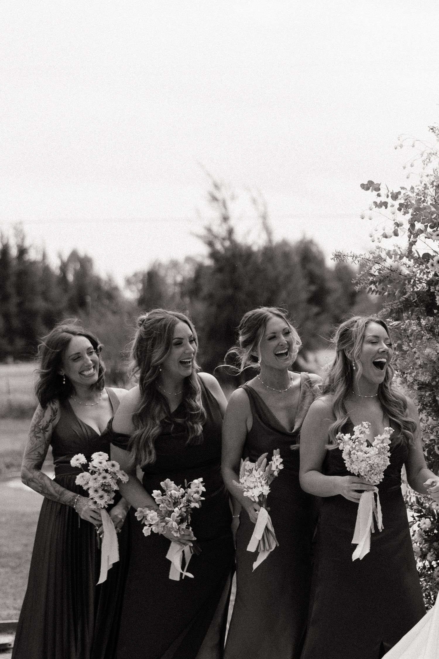 Four bridesmaids in dark gowns laugh together holding white floral bouquets in a black and white photo.