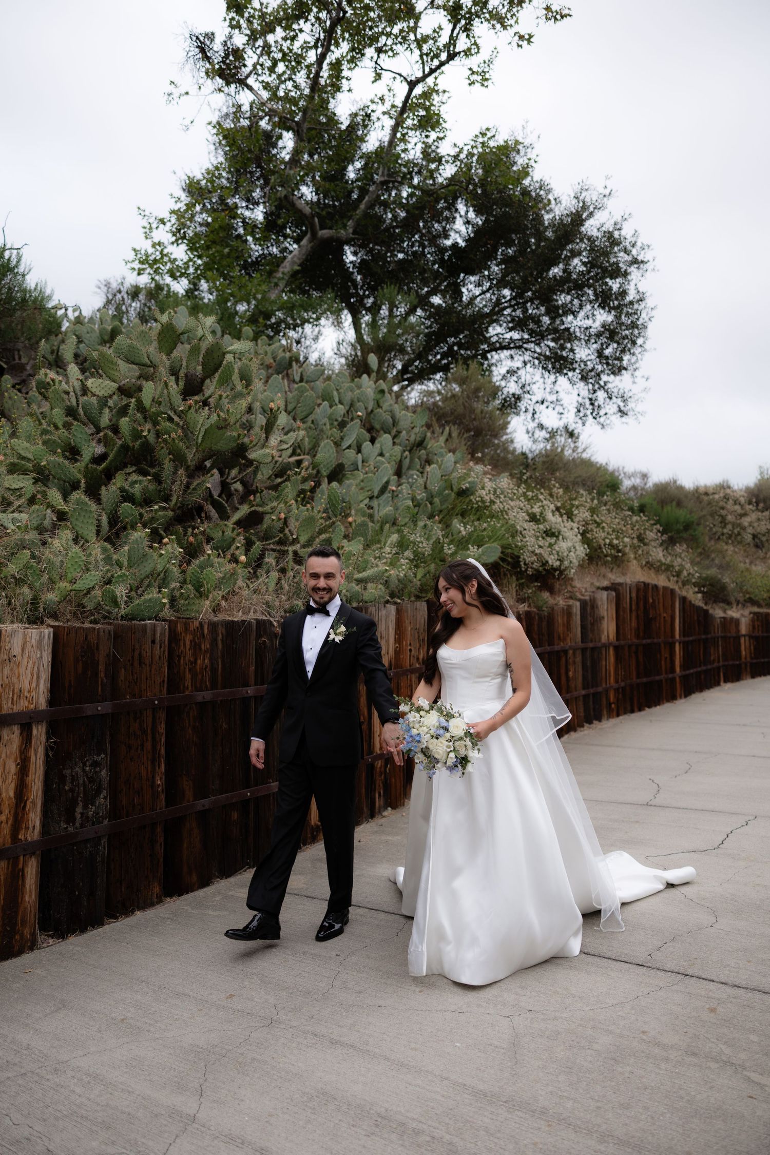 Bride and groom walking hand in hand along a path with cactus and wooden fence backdrop.