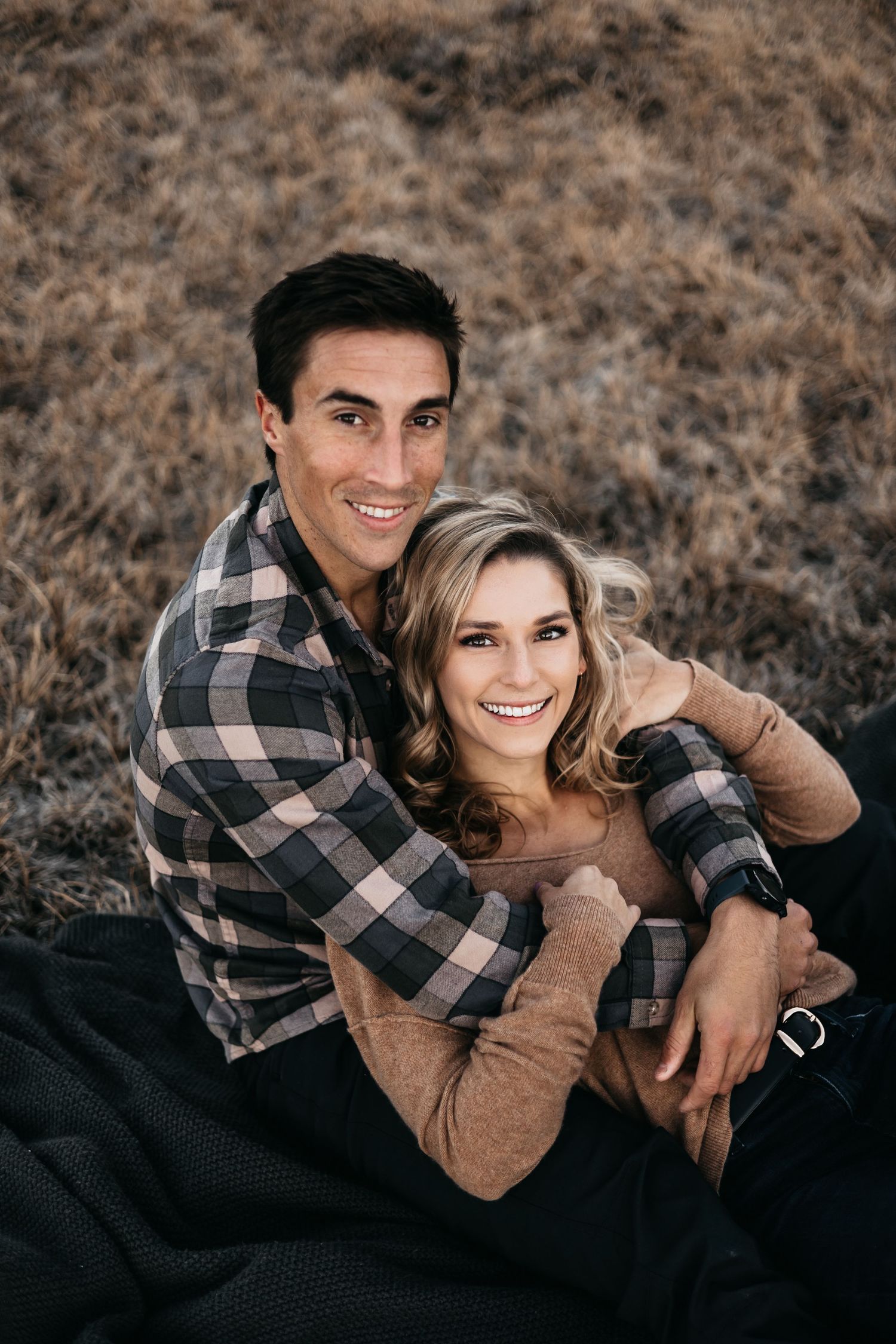 A smiling couple in casual attire embraces warmly while sitting together on a blanket in a dry field.