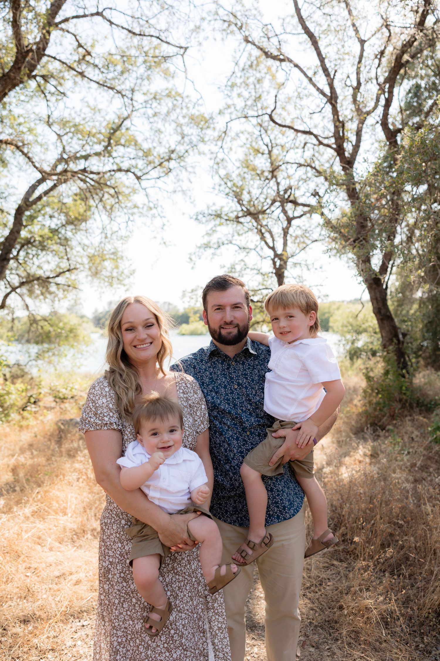 A smiling family of four poses outdoors, parents holding two young boys among trees near a sunlit river.