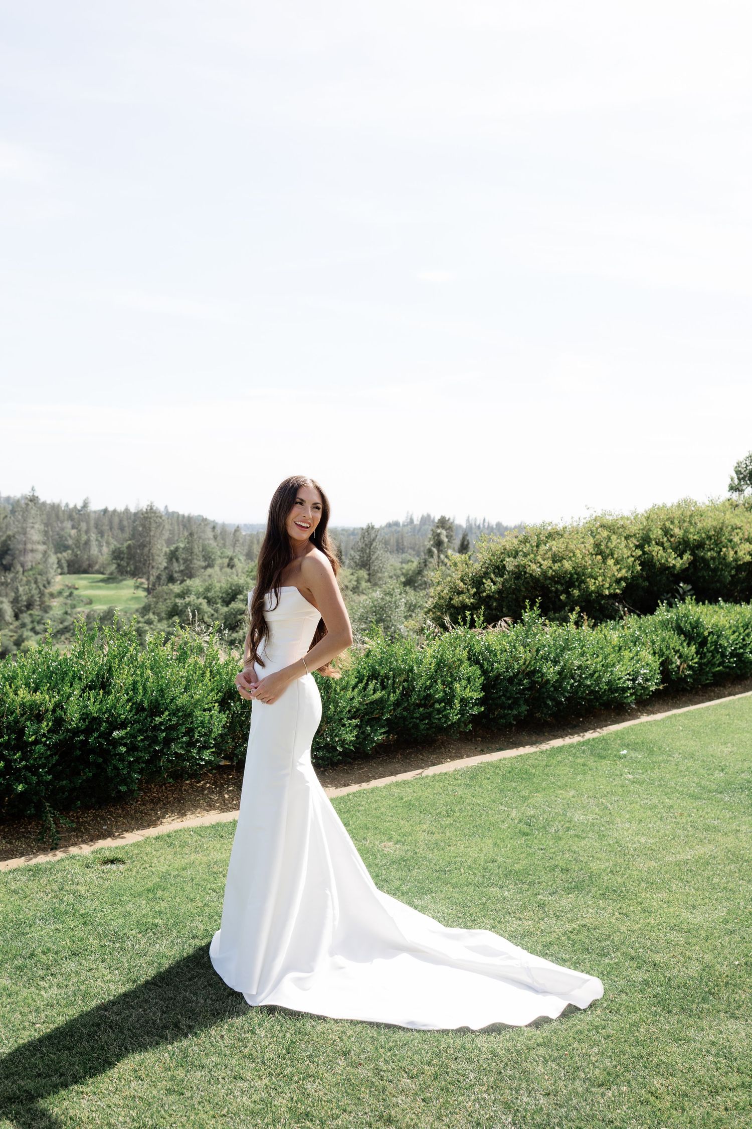 Bride in elegant white mermaid gown with long train standing on manicured lawn.