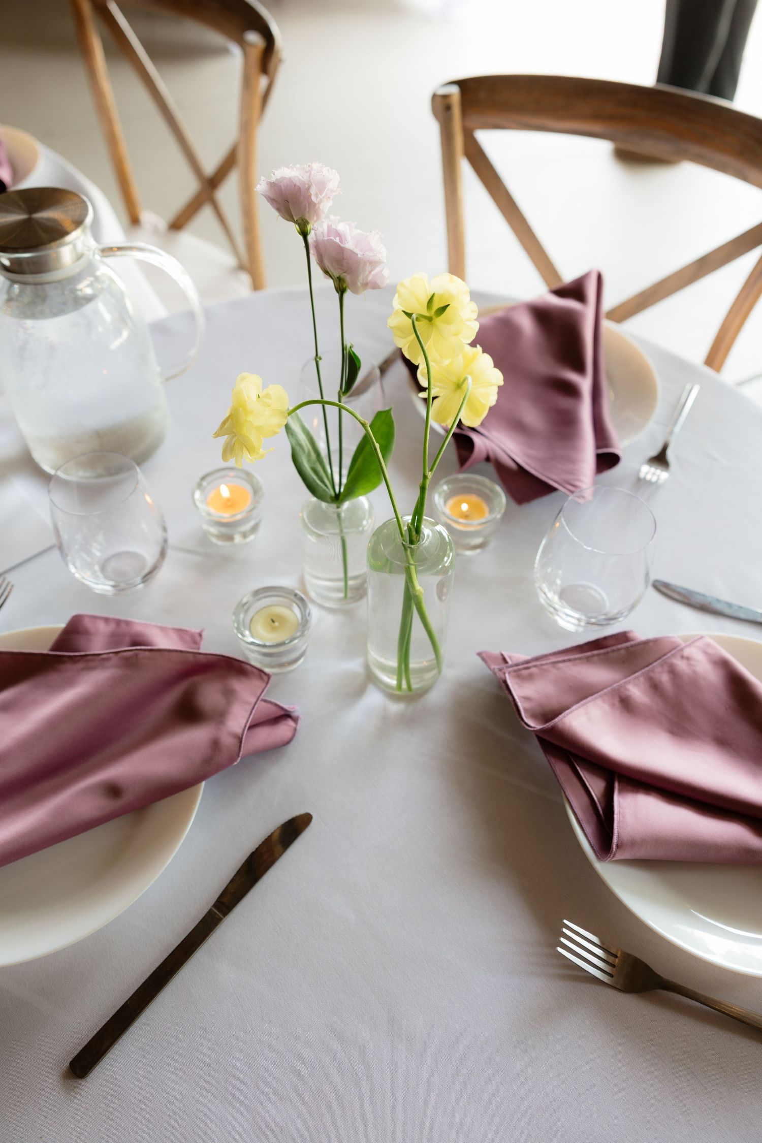 Elegant wedding table setting with dusty rose napkins, white plates, candles, and delicate pastel flowers in glass vases.