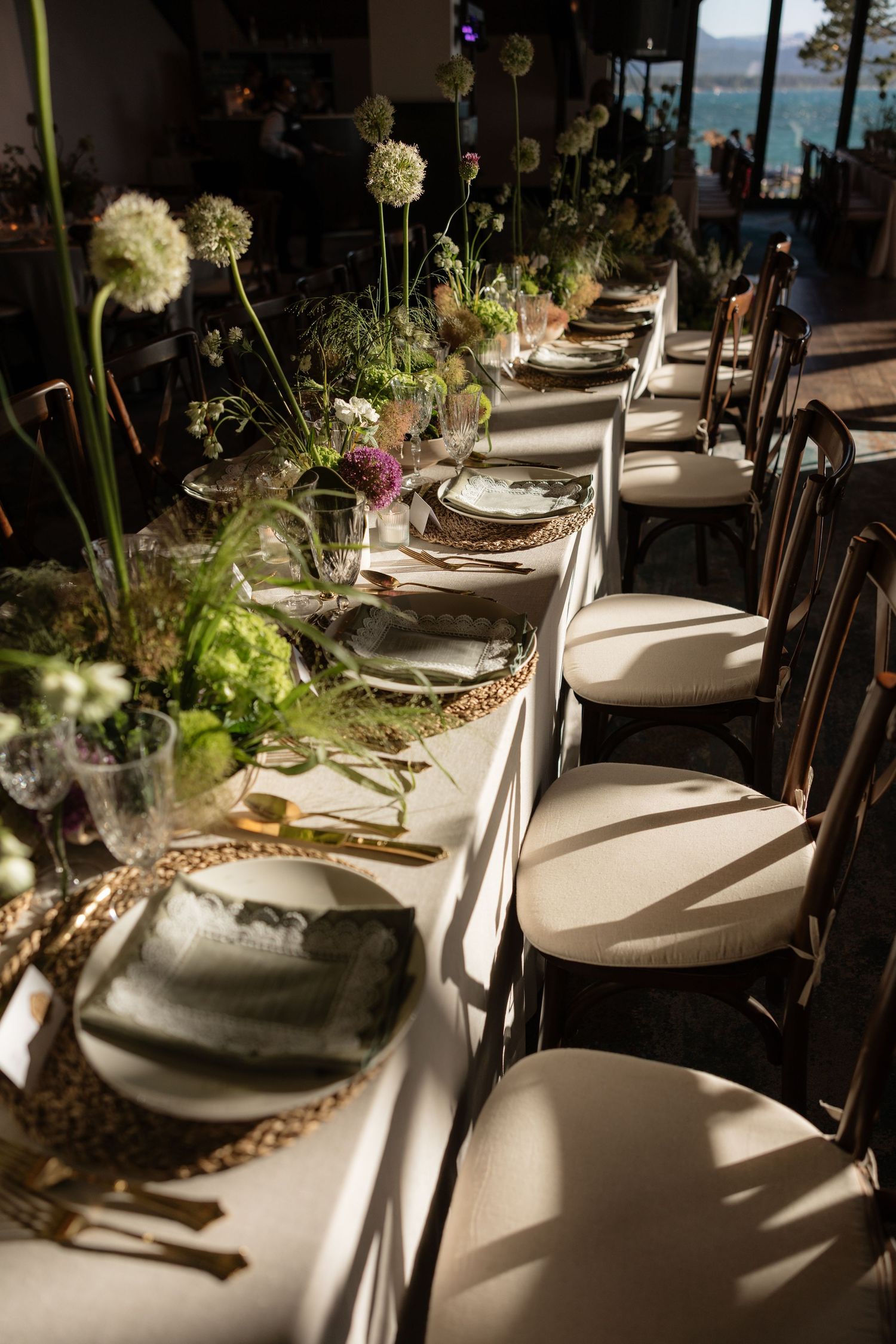 Elegant wedding reception table with white chairs, gold cutlery, woven chargers, and lush floral centerpieces in natural light.