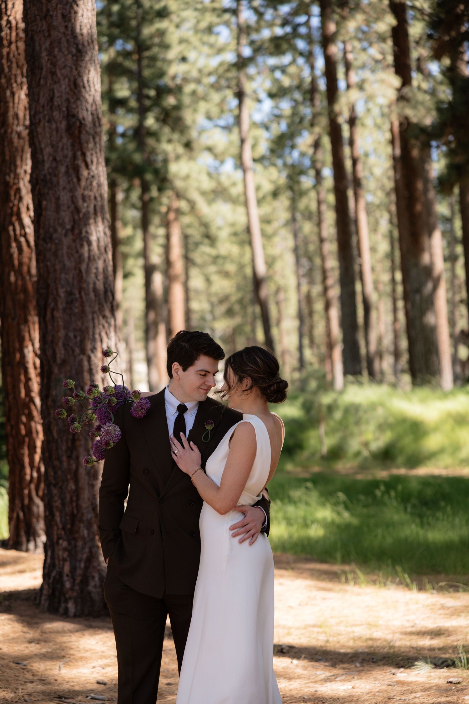 Bride and groom embrace in a sunlit pine forest, wearing a white gown and dark suit with floral bouquet.